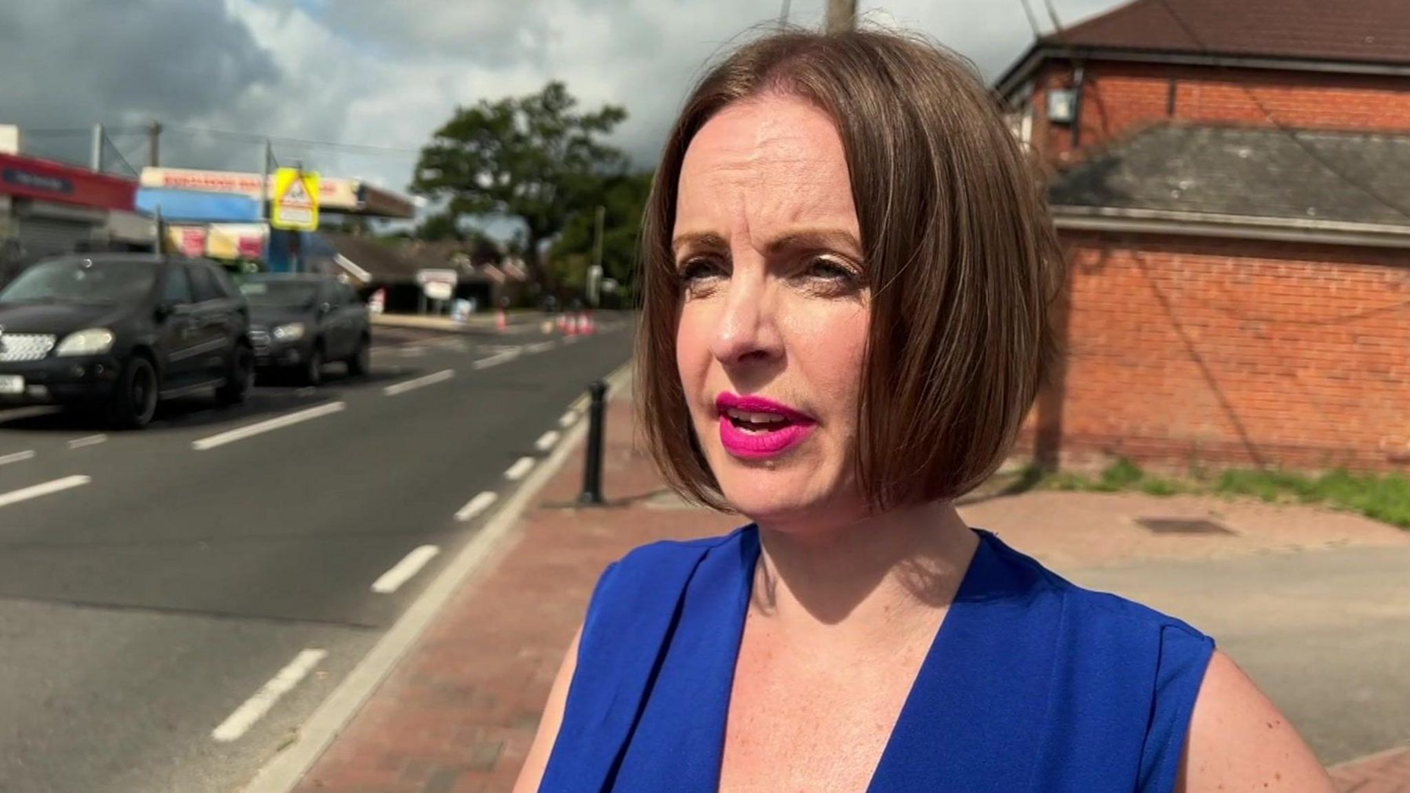 Cheryl Lodwidge standing in the street. She has a brown bob haircut, bright pink lipstick and a royal blue sleeveless top