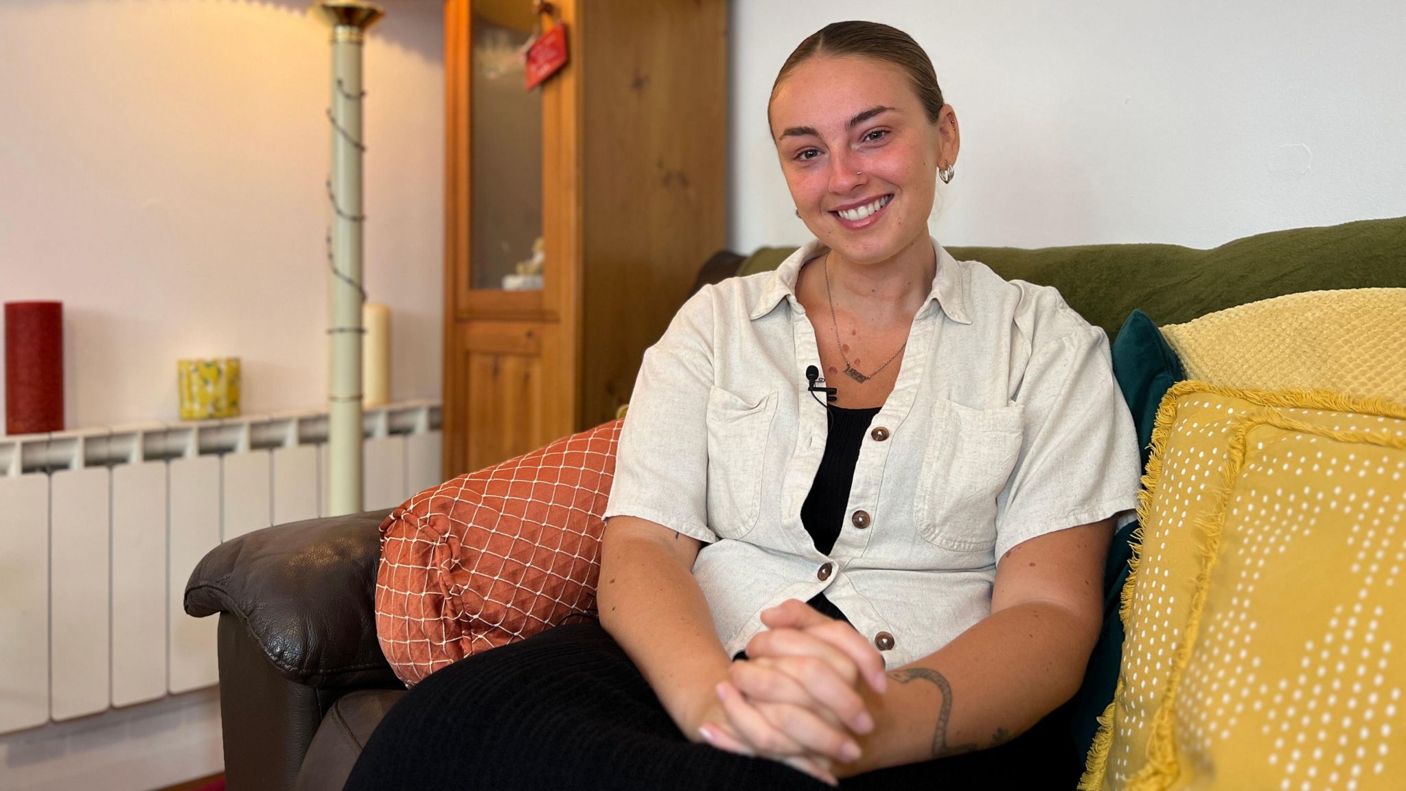 Antonia smiles at the camera as she sits on a sofa with a lamp next to her. Her brown hair is tied bag and she is wearing a beige shirt with her hands clasped together in the foreground.