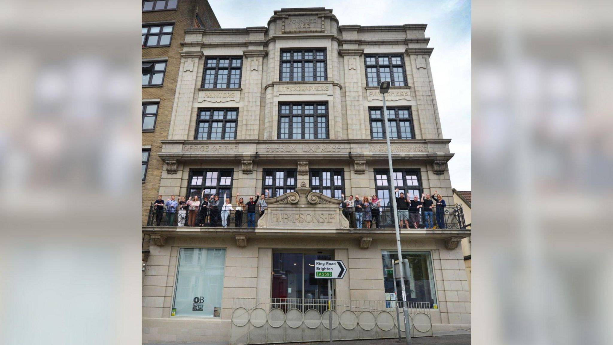 An old building, with four floors. Roughly 25 people are gathered on the balcony of the building, they are cheering