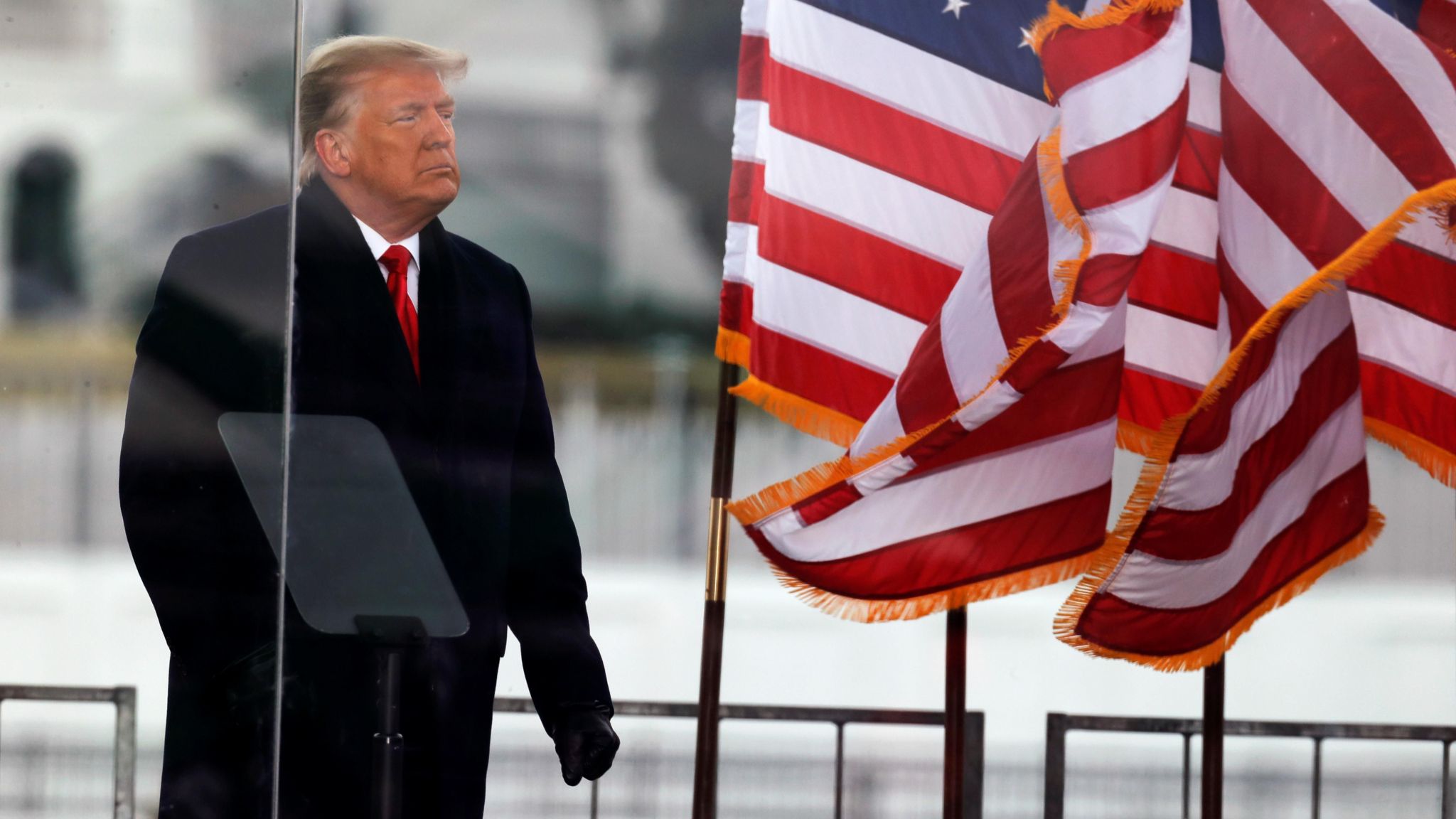 Trump in dark overcoat next to flag behind plexiglass