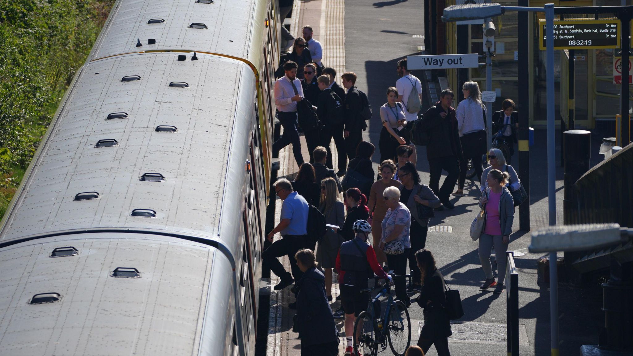 People get on and off a Merseyrail train on the Southport line. They are concentrated around the doors of the train. In the background there is a live electronic board detailing the train's arrival and stops, and there is also a way out sign directing passengers off the platform.