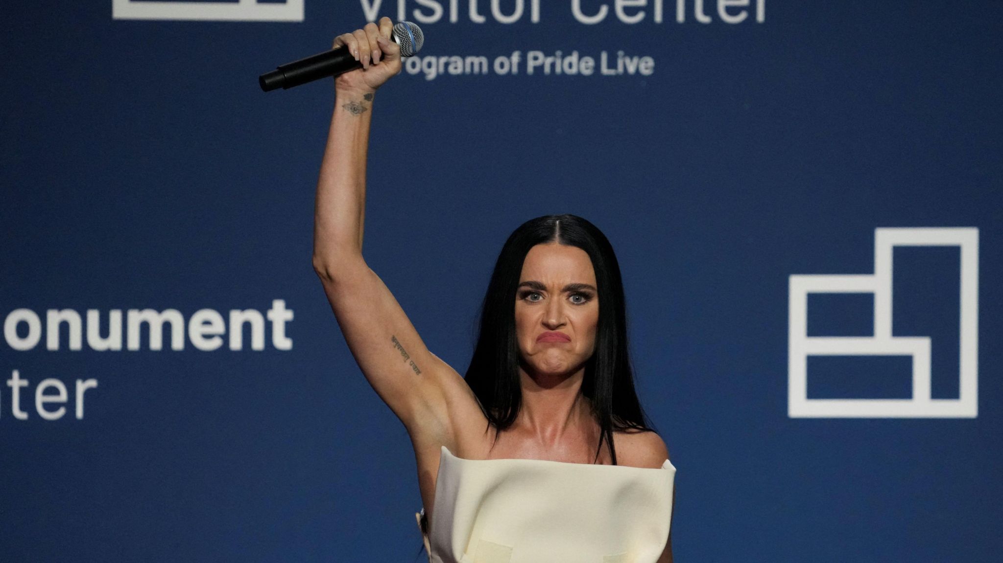 Katy Perry dressed in white dress raises fist in the air while holding a microphone