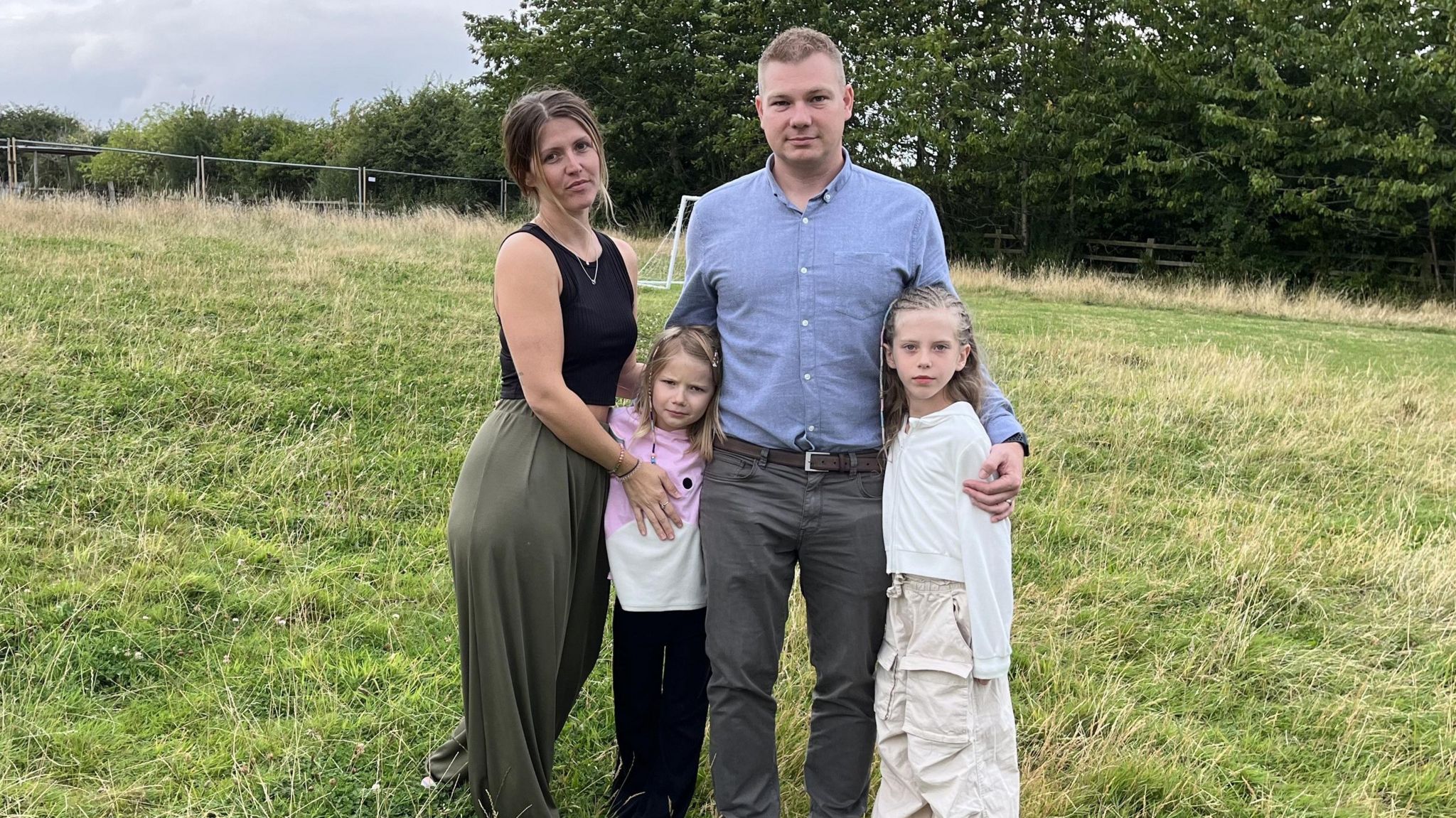 A family standing on a green field. On the left is the mother, who is holding a child to her side. In the middle is the father, Anthony, who is in a blue shirt. To his right is another child, who he has his arm around.