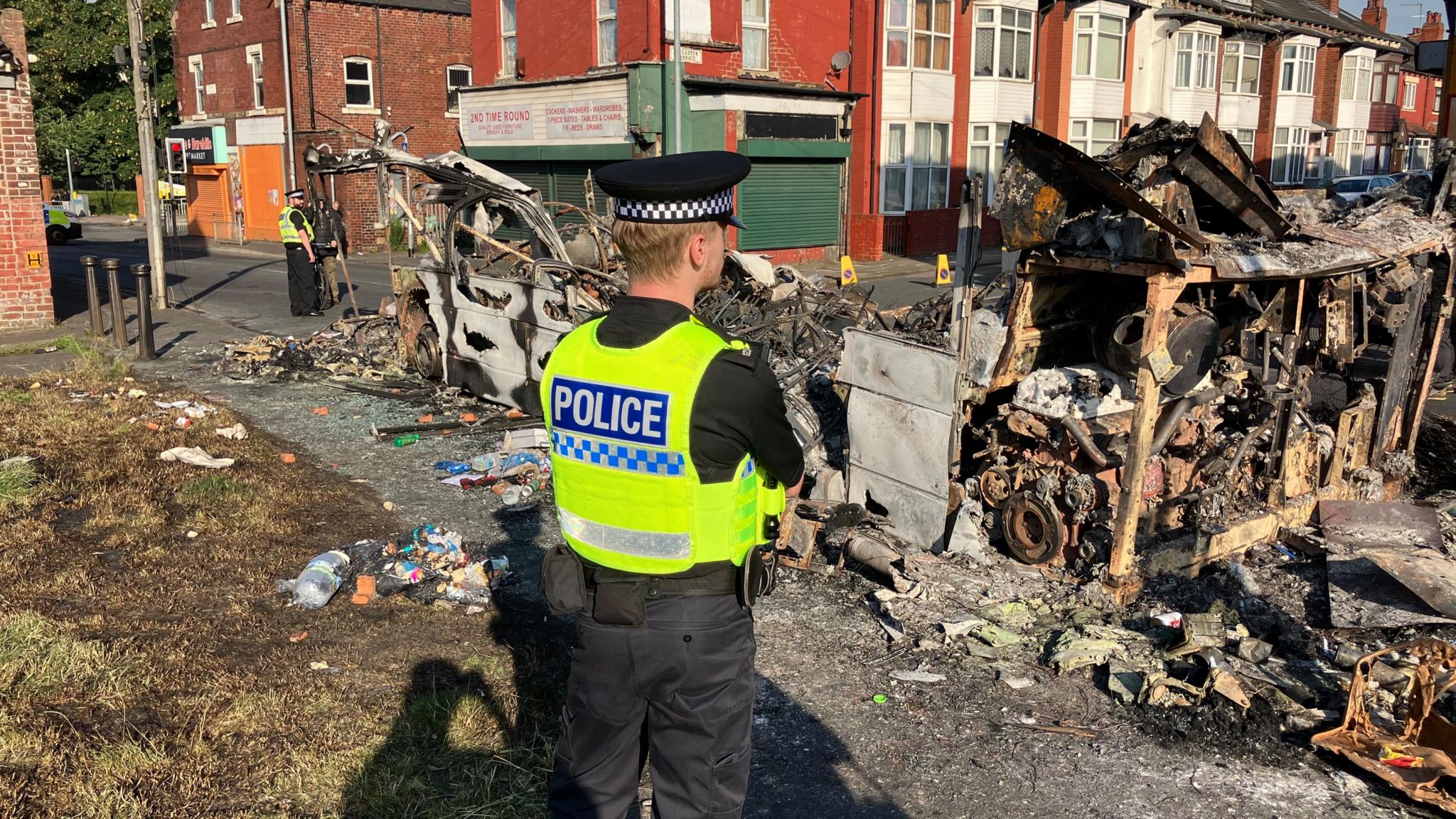 A police officer stands in front of a burnt-out bus