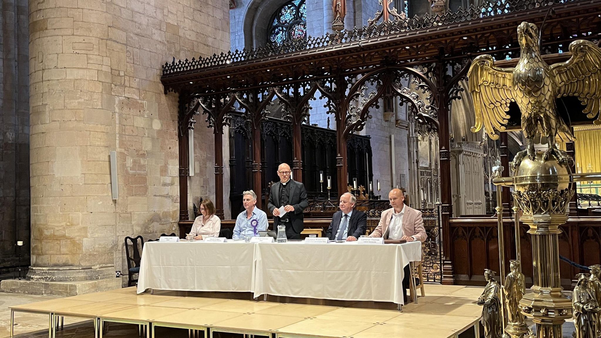 Four people sitting either side of a standing vicar on a stage in Tewskesbury Abbey next to a large golden eagle lectern