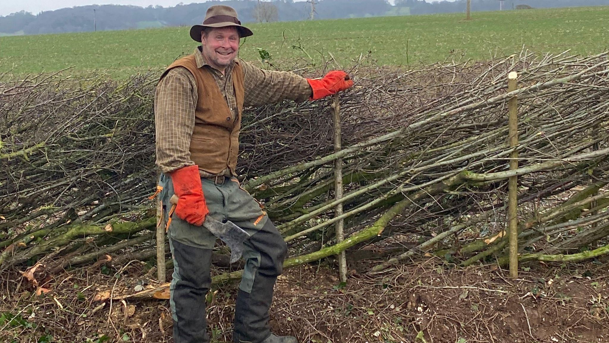 A smiling Tom Vowles stands in a field holding a large cutting tool in one hand and rests the other hand on the top of a cut branch that now forms a post in a weaved fence. He is wearing a brown checked shirt, brown waist coast and green and black trousers, as well as sturdy bright red gloves and a brimmed hat