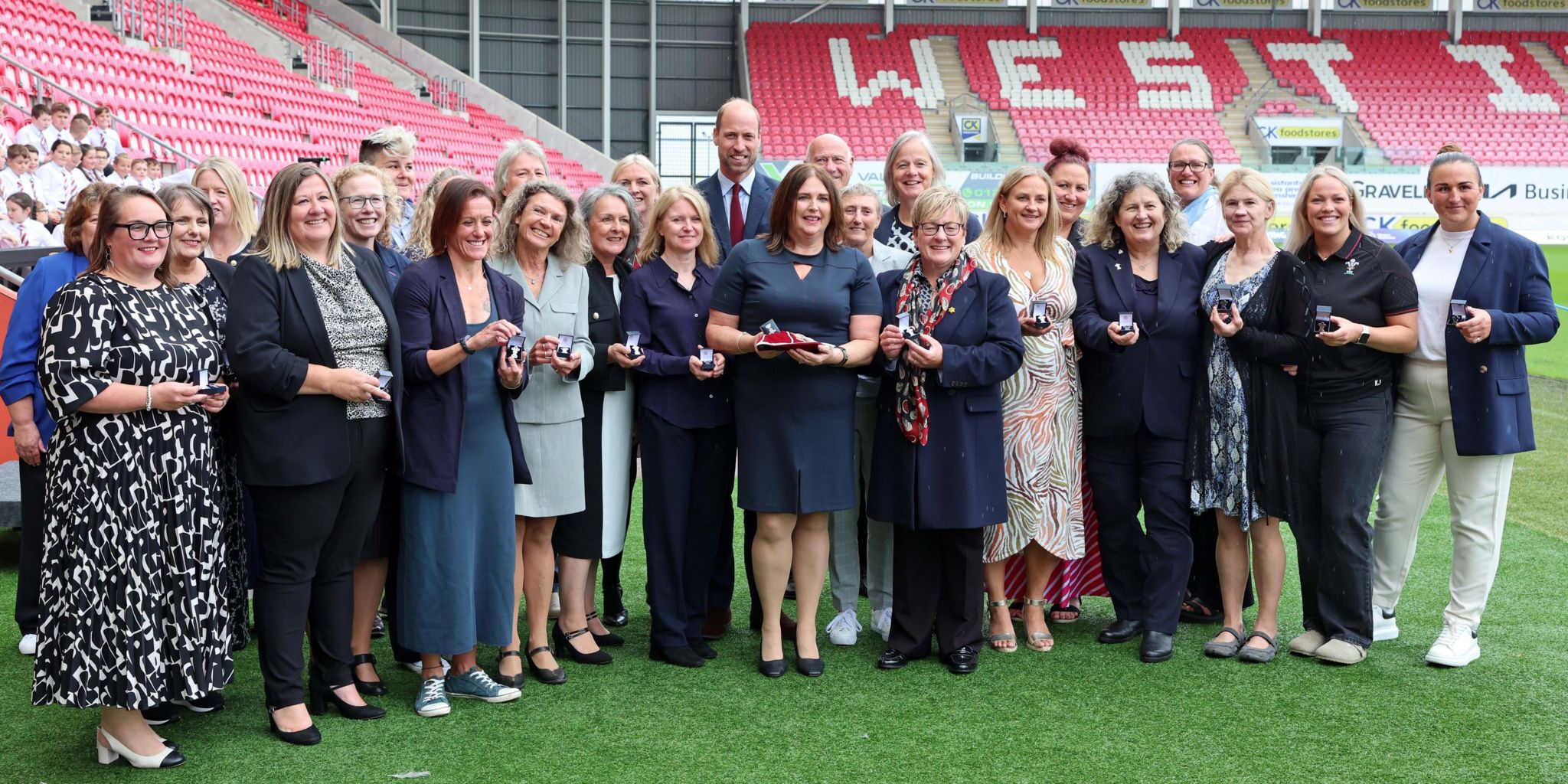 Picture of Prince William with a number of the Welsh women's rugby team, smiling and holding their caps at the Parc Y Scarlets stadium. 