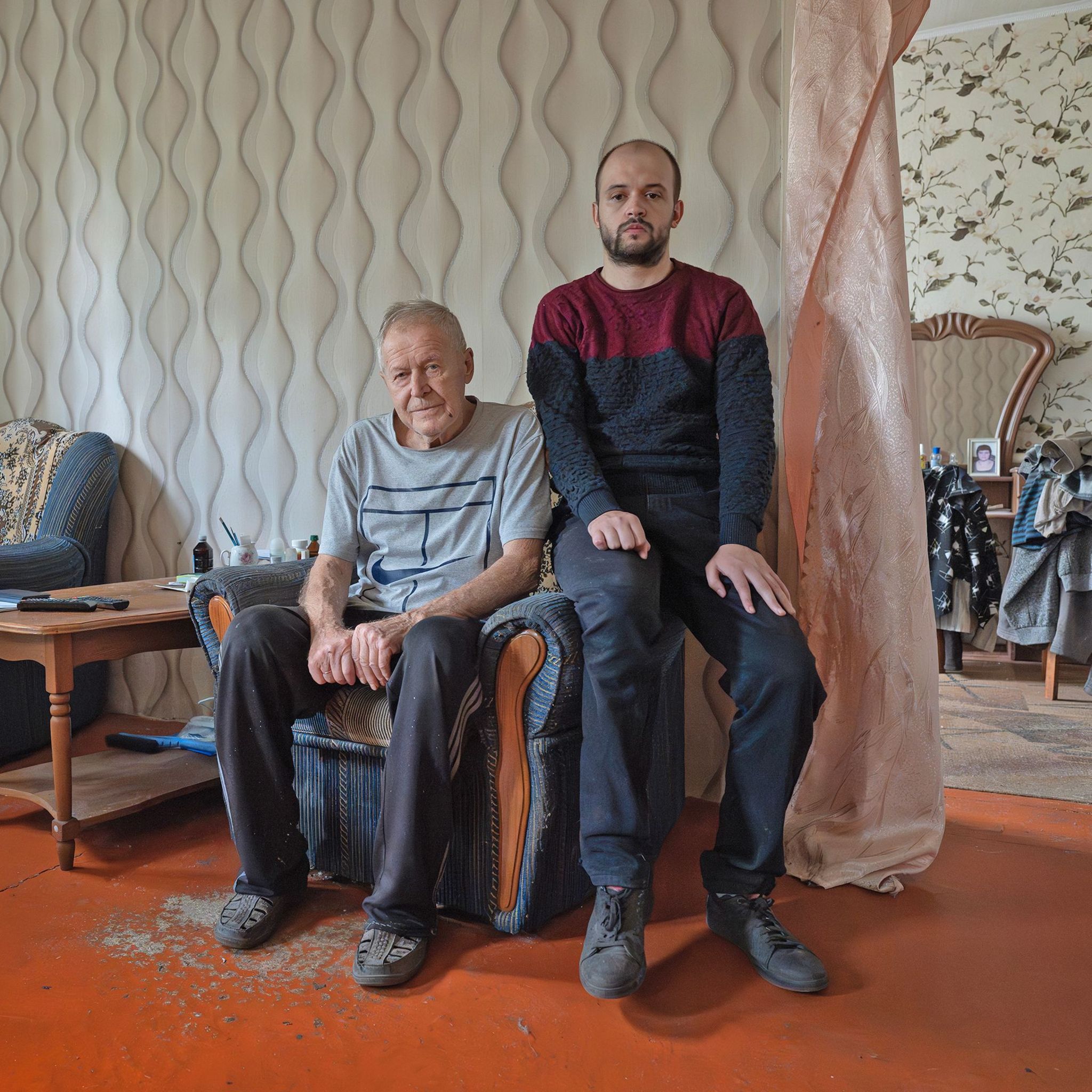 Tetyana's husband and adult disabled son sit on a chair at their home, with piles of clothes visible in the background