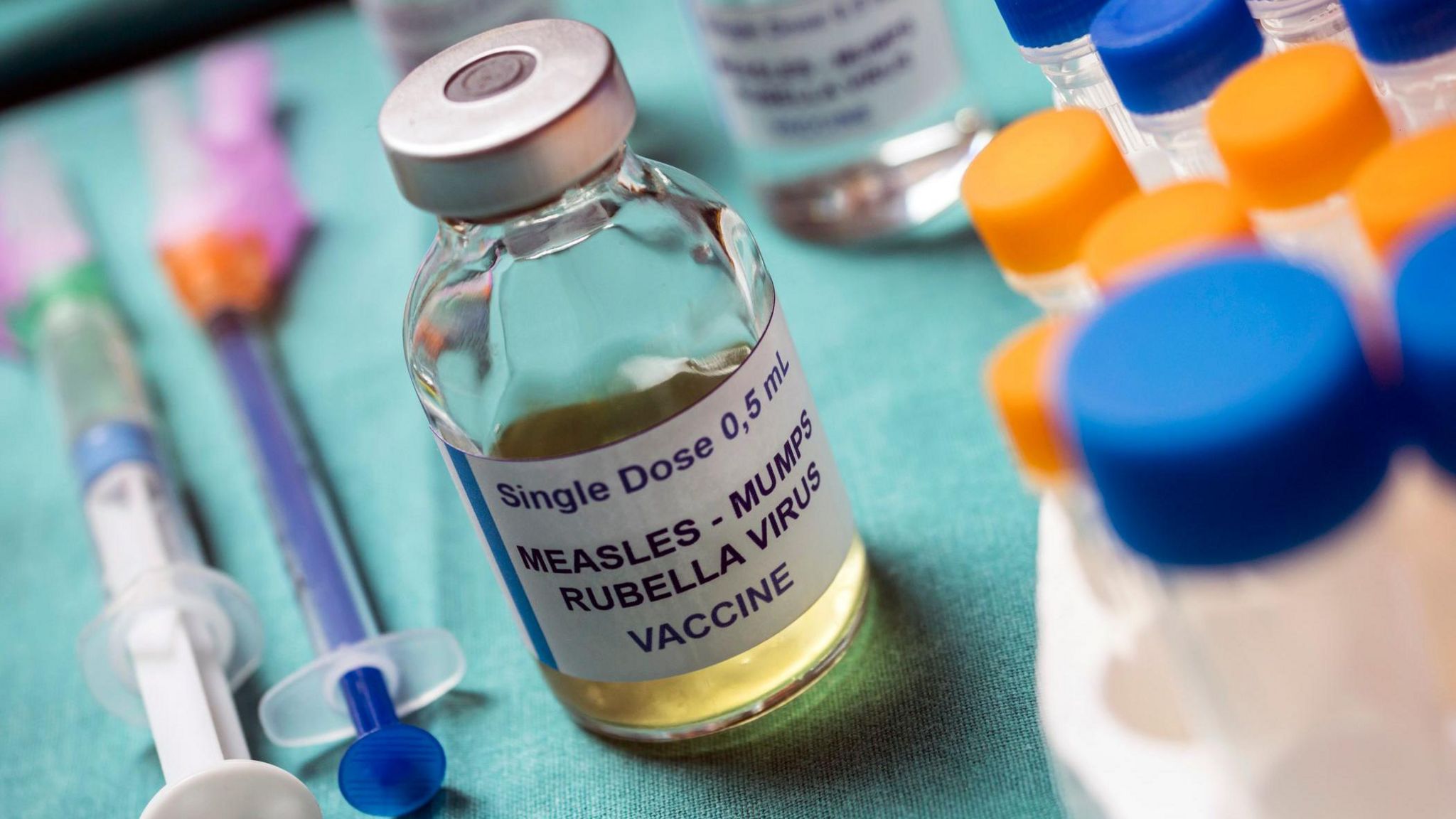 A vile of measles mumps and rubella vaccine sat on a green cloth beside needles and other medical equipment