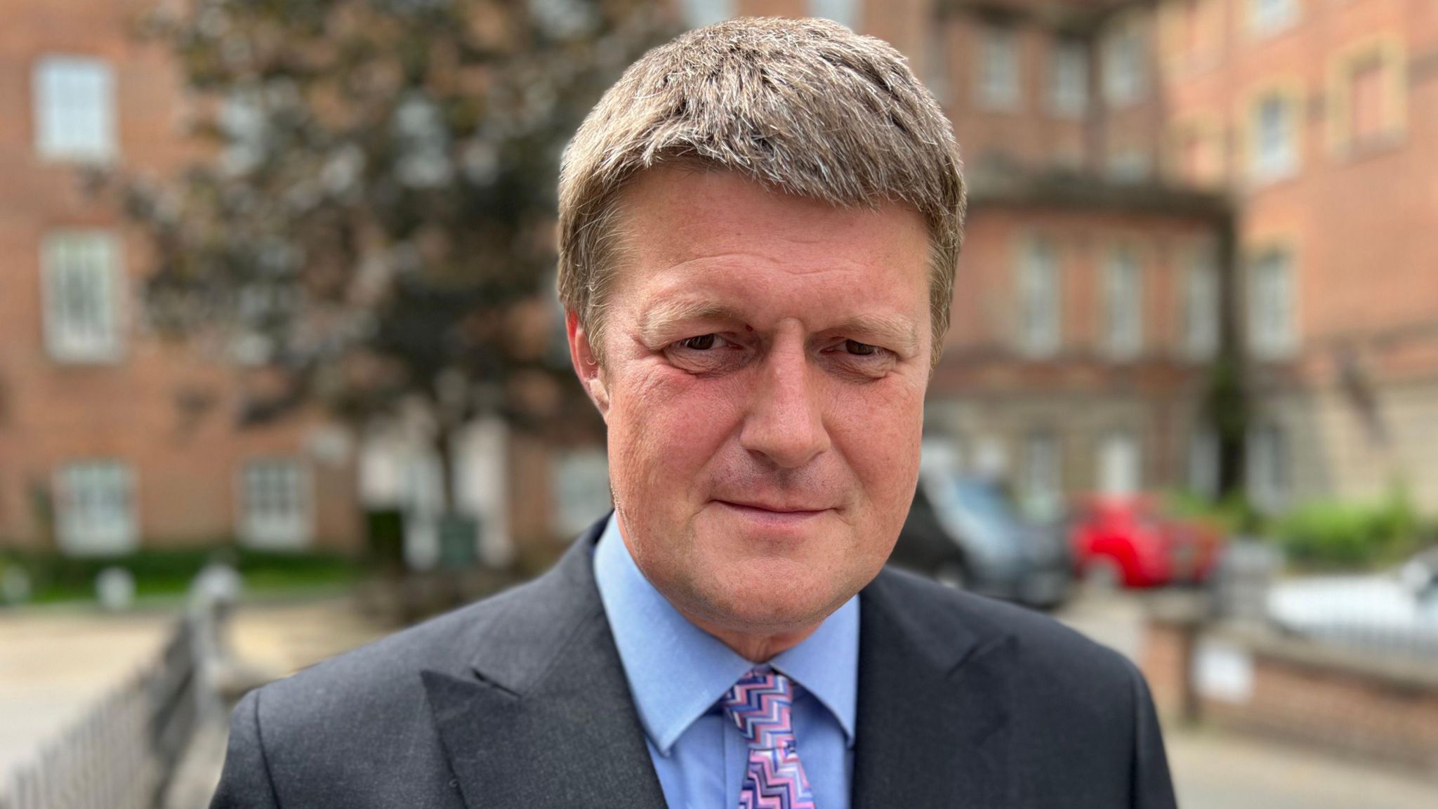 Head and shoulders image of Richard Clewer in a dark suit with a blue shirt and pink, zig, zag pattern tie