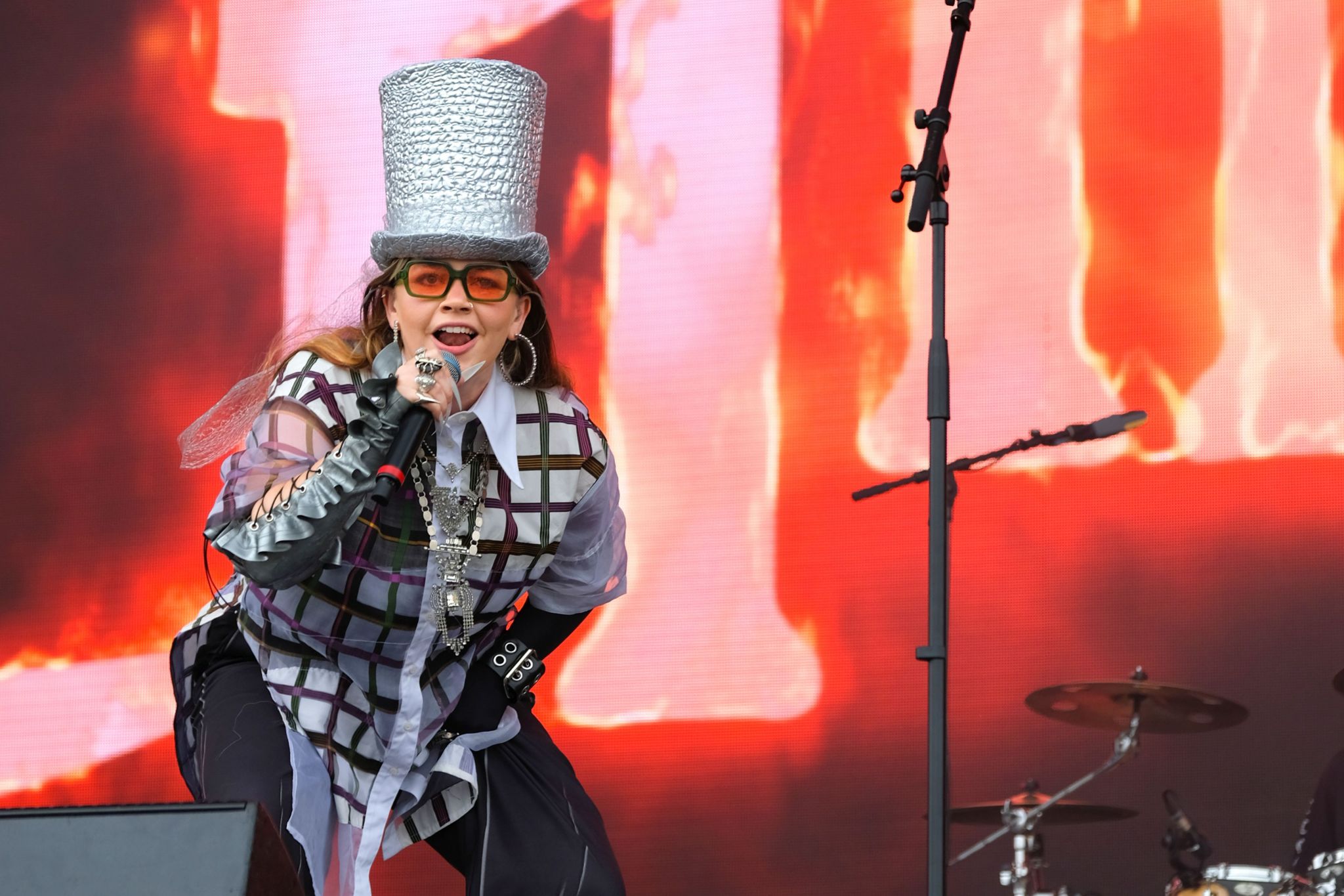 Pop artist CHINCHILLA performs wearing a silver top hat, a checked shirt and orange tinted sunglasses. She also wears black trousers whilst holding a microphone, singing on stage.