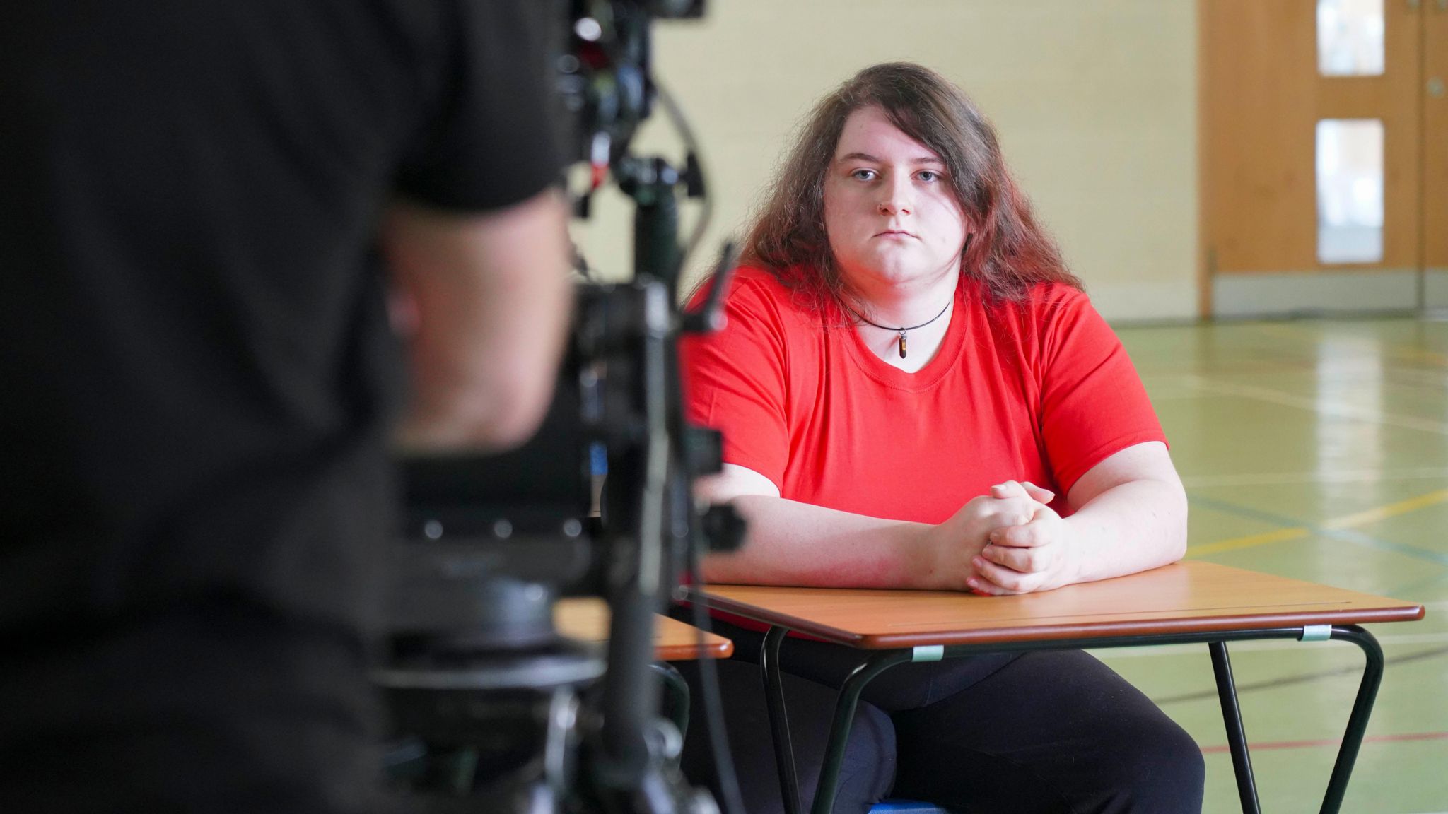 A young woman in a red top sitting at a desk in a gym hall with her hands clasped together, she is being filmed by a camera