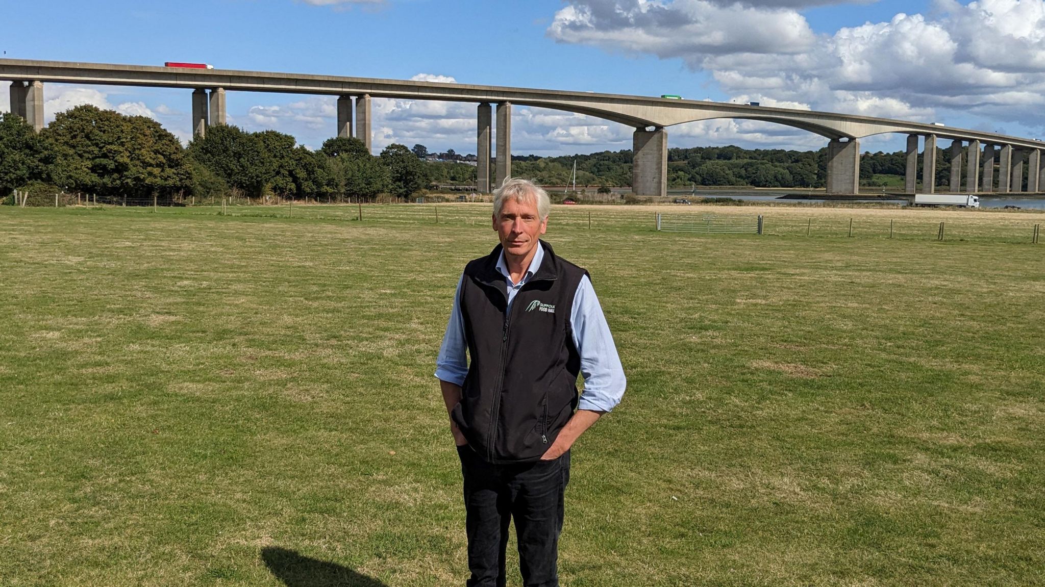 Oliver Paul, standing on grass, with the River Orwell and the Orwell Bridge behind him