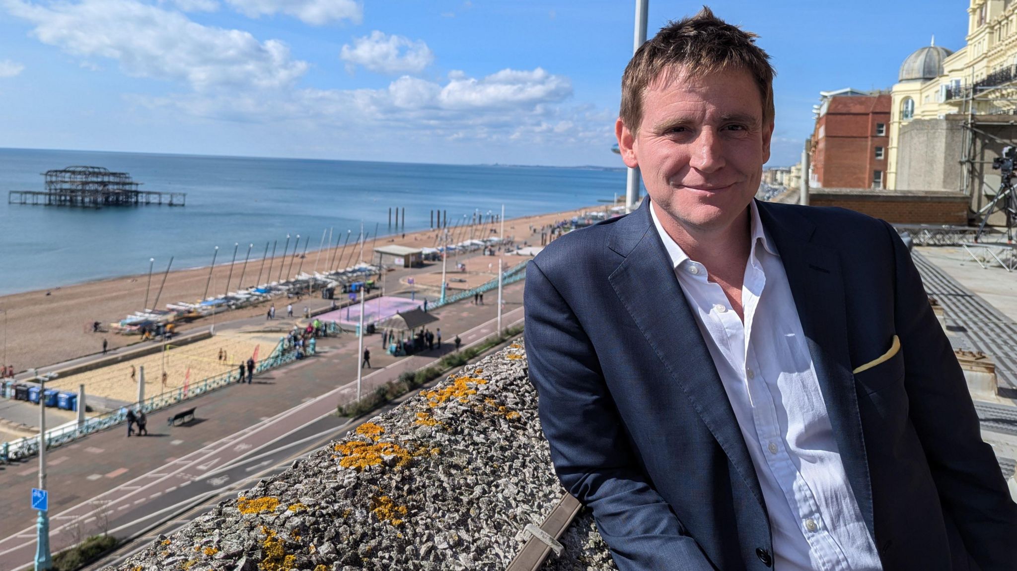 Mike Martin MP leans against a balcony overlooking Brighton Beach and Brighton Pier on a sunny day. He's wearing a navy suit, a pale open-necked shirt and his short, brown hair is spiked up a little in the middle 