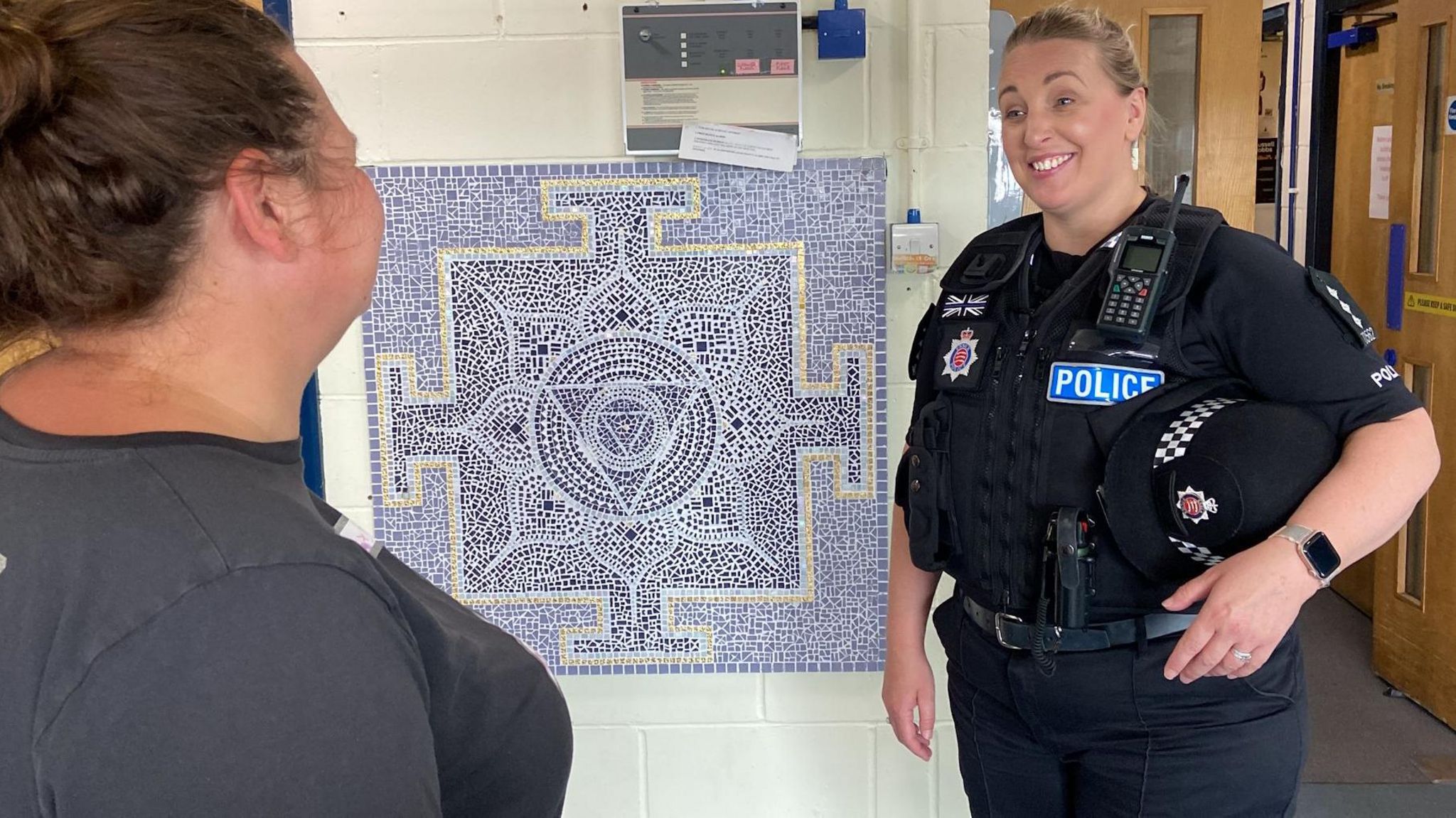 Tendring district commander Chief Inspector Ella Latham in police uniform, smiling and looking at another woman while standing by a blue mosaic on a wall