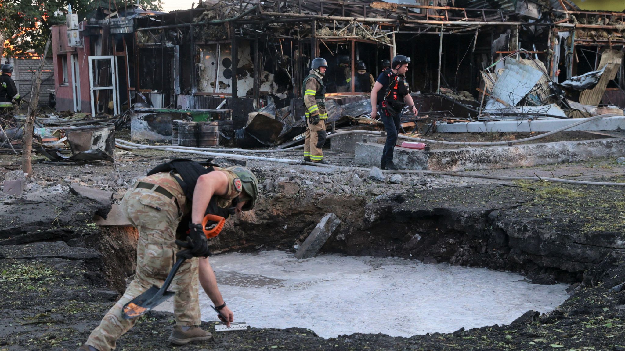 A Ukrainian official inspects a shell crater in Vilniansk, Zaporizhzhia region, on 29 June 2024, as firemen work on nearby burned-out buildings.