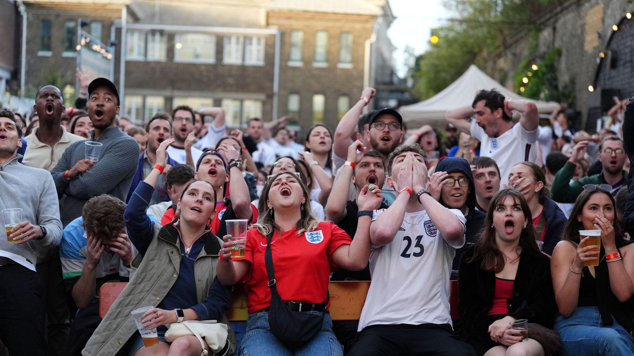 A group of England fans sitting in a fan zone. The are reacting to a moment in the game, with some people with their mouths open, others with hands on their heads, and others covering their mouths