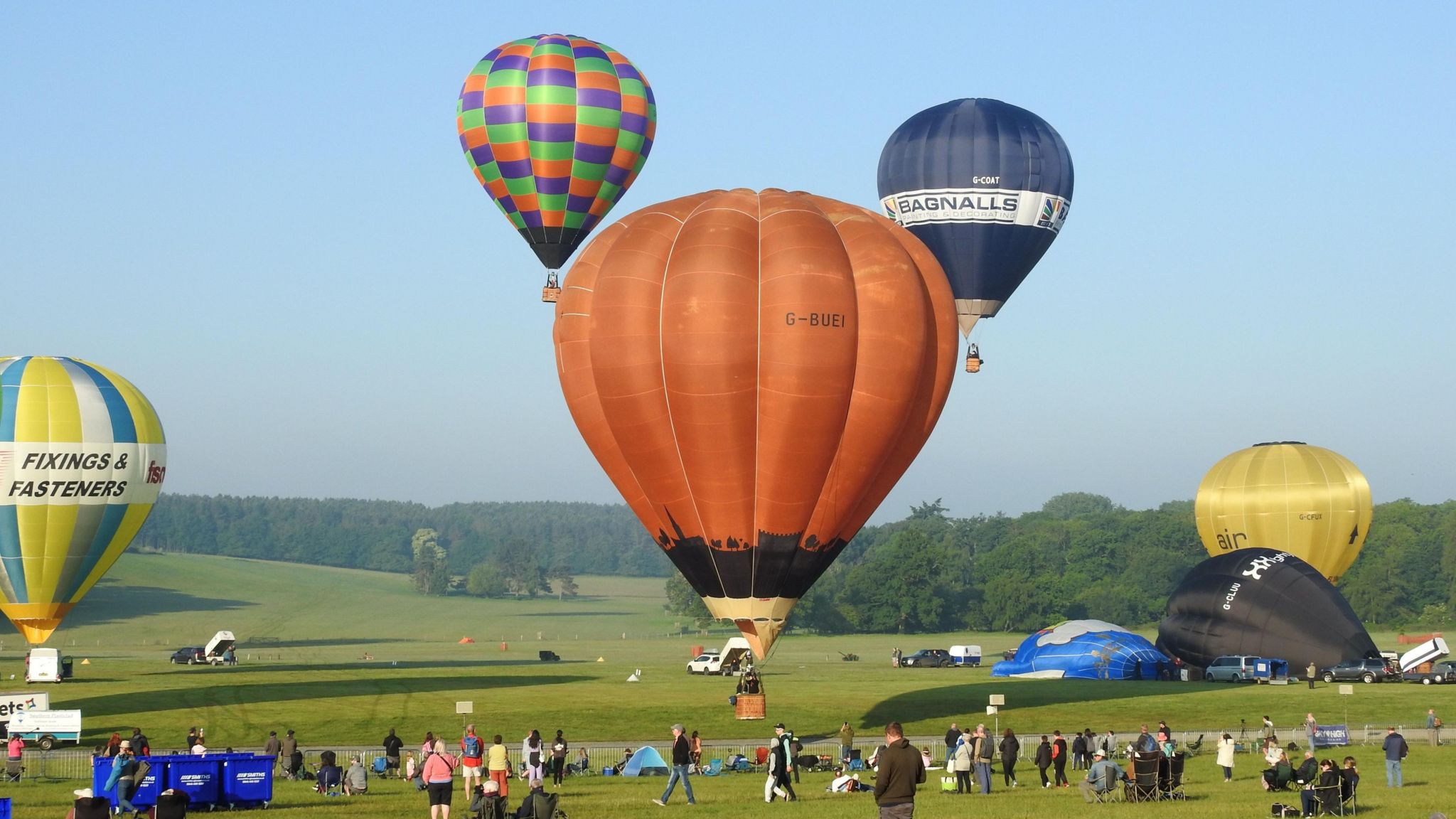 A field with hot balloons taking off and a number of people standing on the ground watching