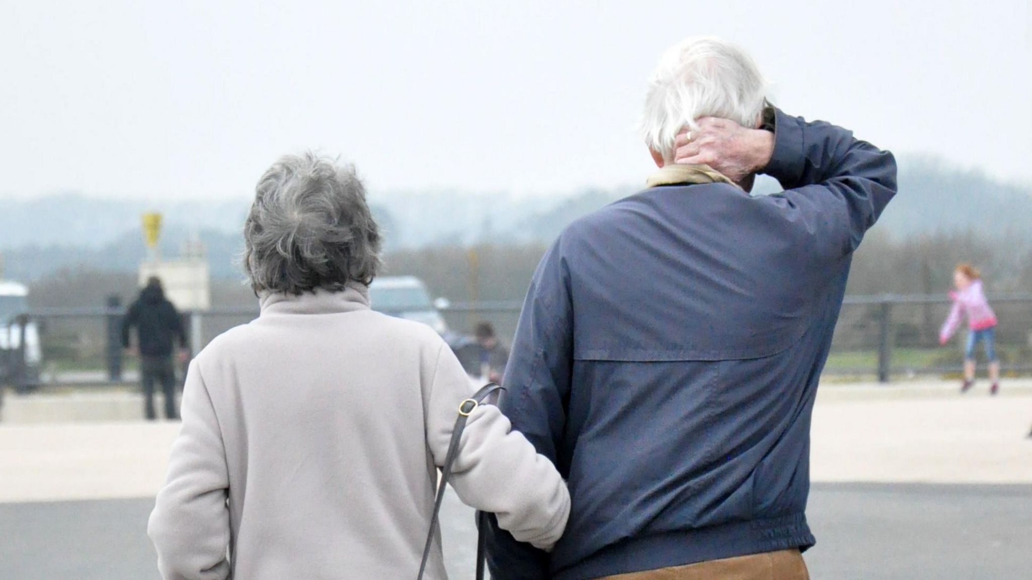 Two elderly people walking side by side. One on the left in a grey jumper, the one of the right in a blue jumper. Both are walking away.