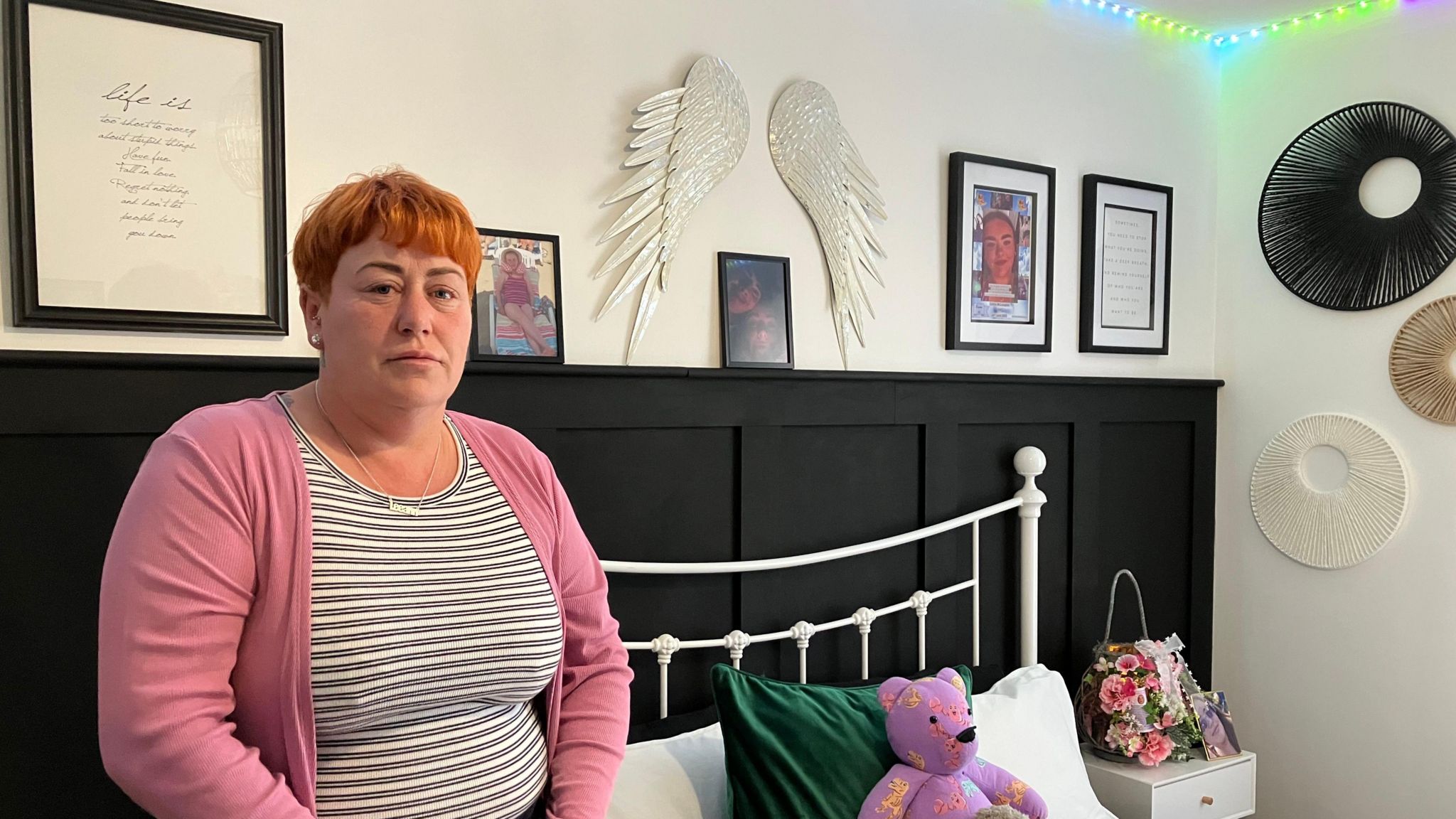 leanne mclaughlin stands beside her daughter caitlin's bed, on which there sits a teddy bear, and beside which is a bunch of flower. a pair of angel wings hang above the bed