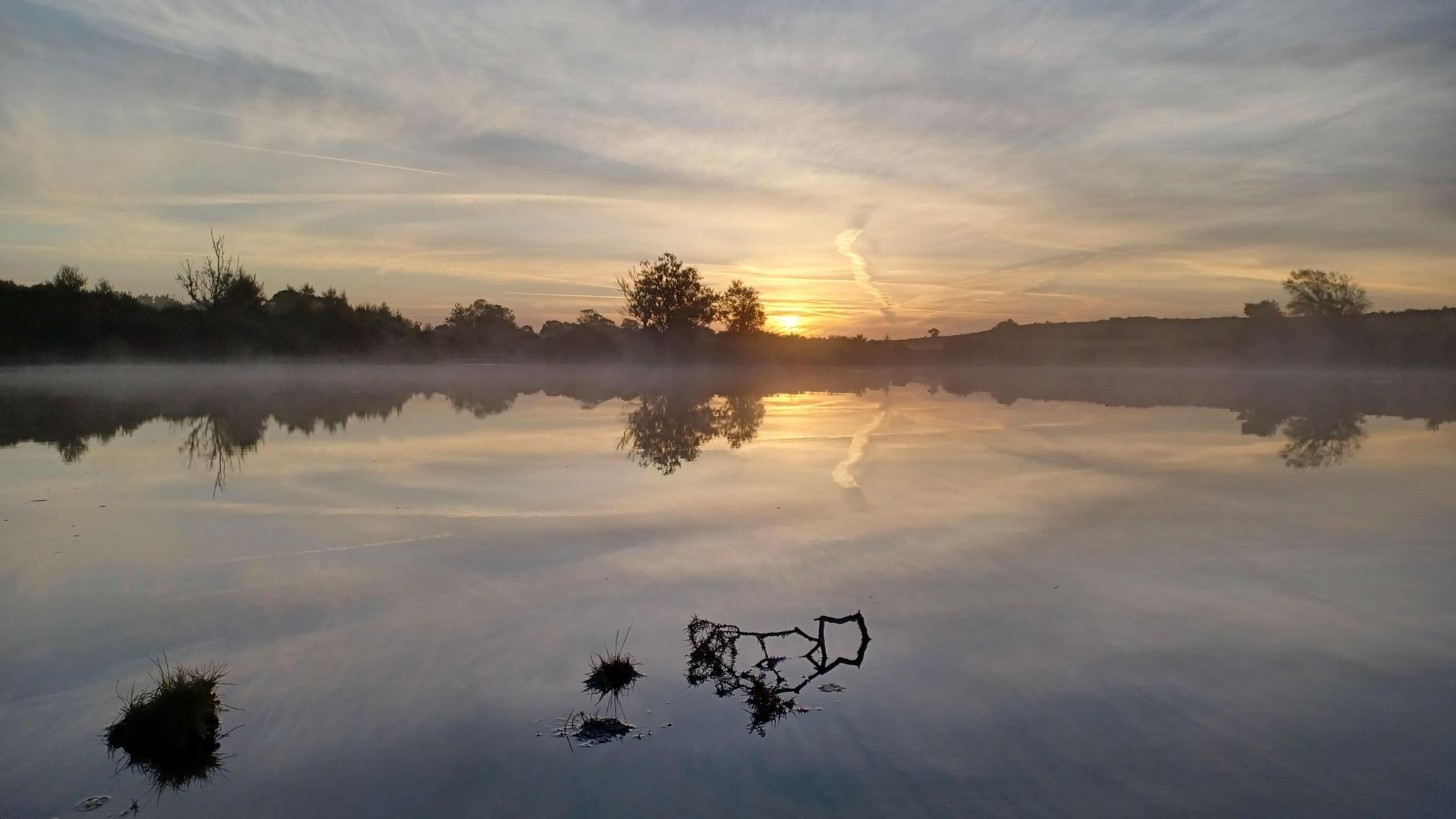 The sunrises over a pond in the New Forest, there is a mist over the water and trees on the far bank that are reflected in the water. The sun is glowing yellow as it appears over the horizon