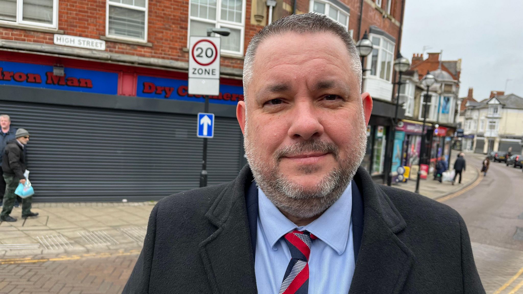 A man in a navy coat, a blue shirt and a red-and-grey tie standing on a high street