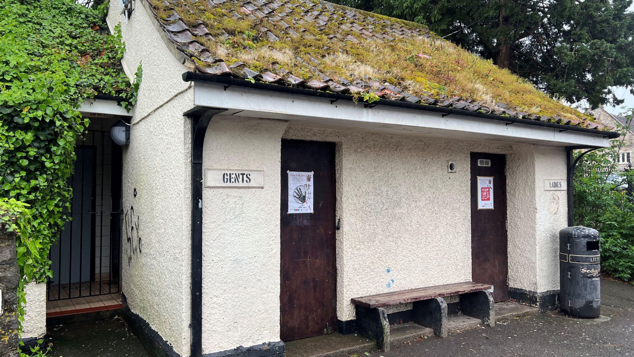 Old toilet block which is closed and locked up. It has vegetation growing on its roof and its doors and gutters are painted black