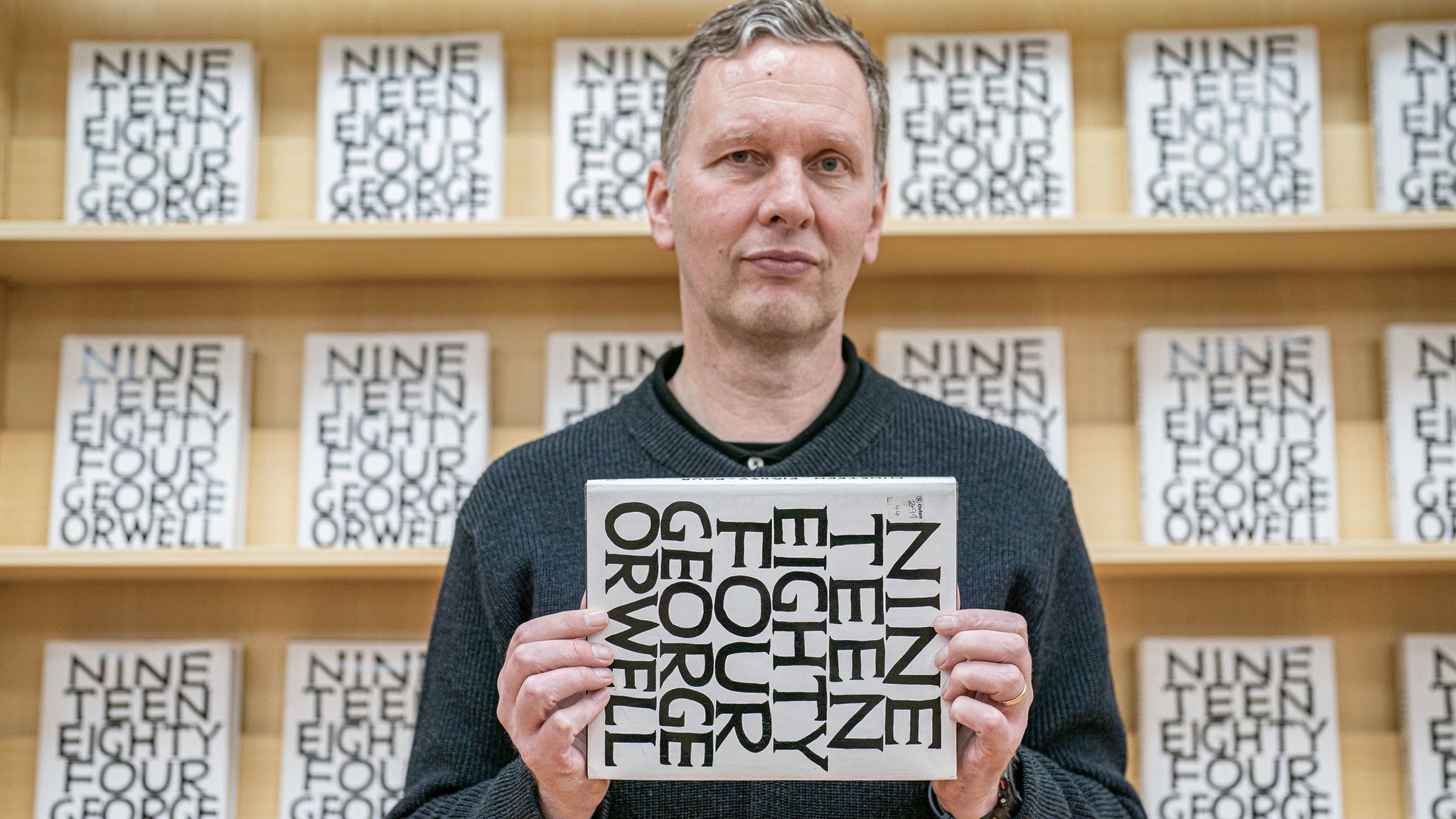 David Shrigley standing in front of a book shelf of his 1984 edition.