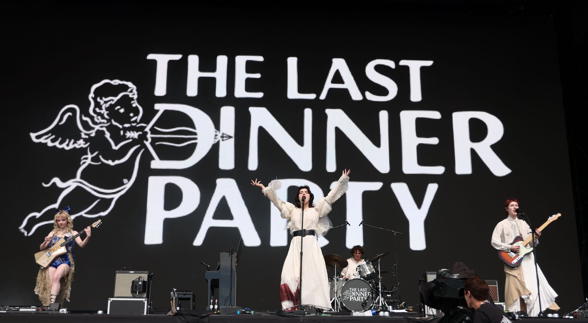 Band members on stage wearing white address a black background with the words 'the Last Dinner Party' written on