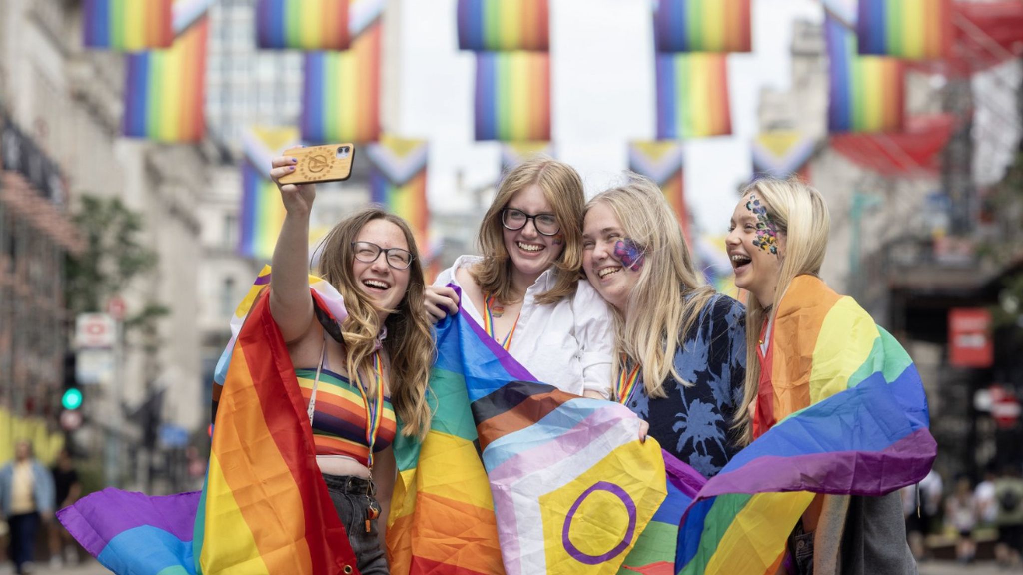 Isabel Cato, Ashleigh Tory, Charley Cook and Matilda Beavers take a selfie on Regent Street in front of Intersex-Inclusive Pride flags which were designed by Valentino Vecchietti