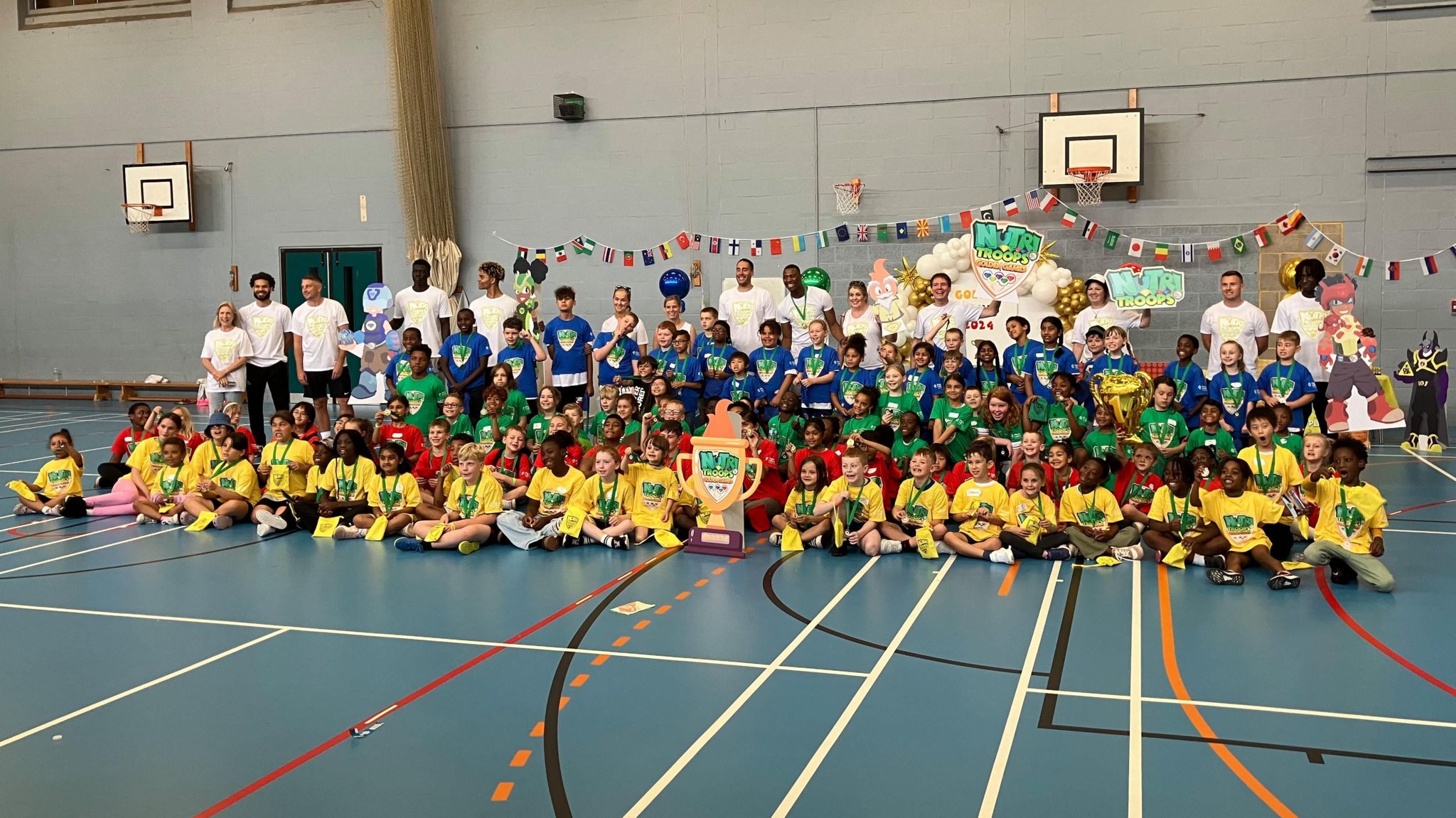 A large crowd of children and some adults in coloured tshirts sitting in a sports hall with flags arranged behind