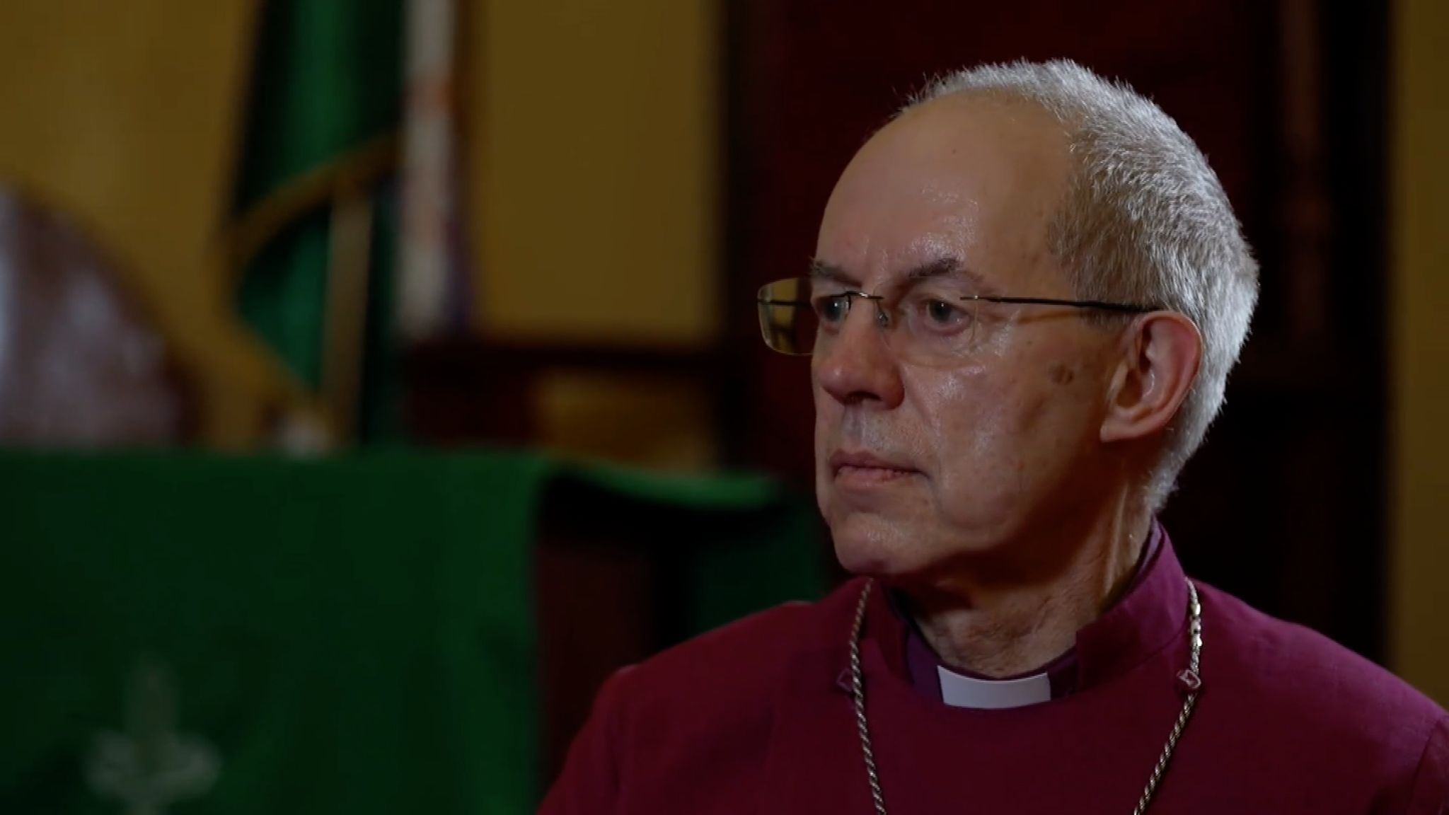Justin Welby, with short balding grey hair and glasses looks to the side in an interview as he wears a purple cassoc and a large cross around his neck.