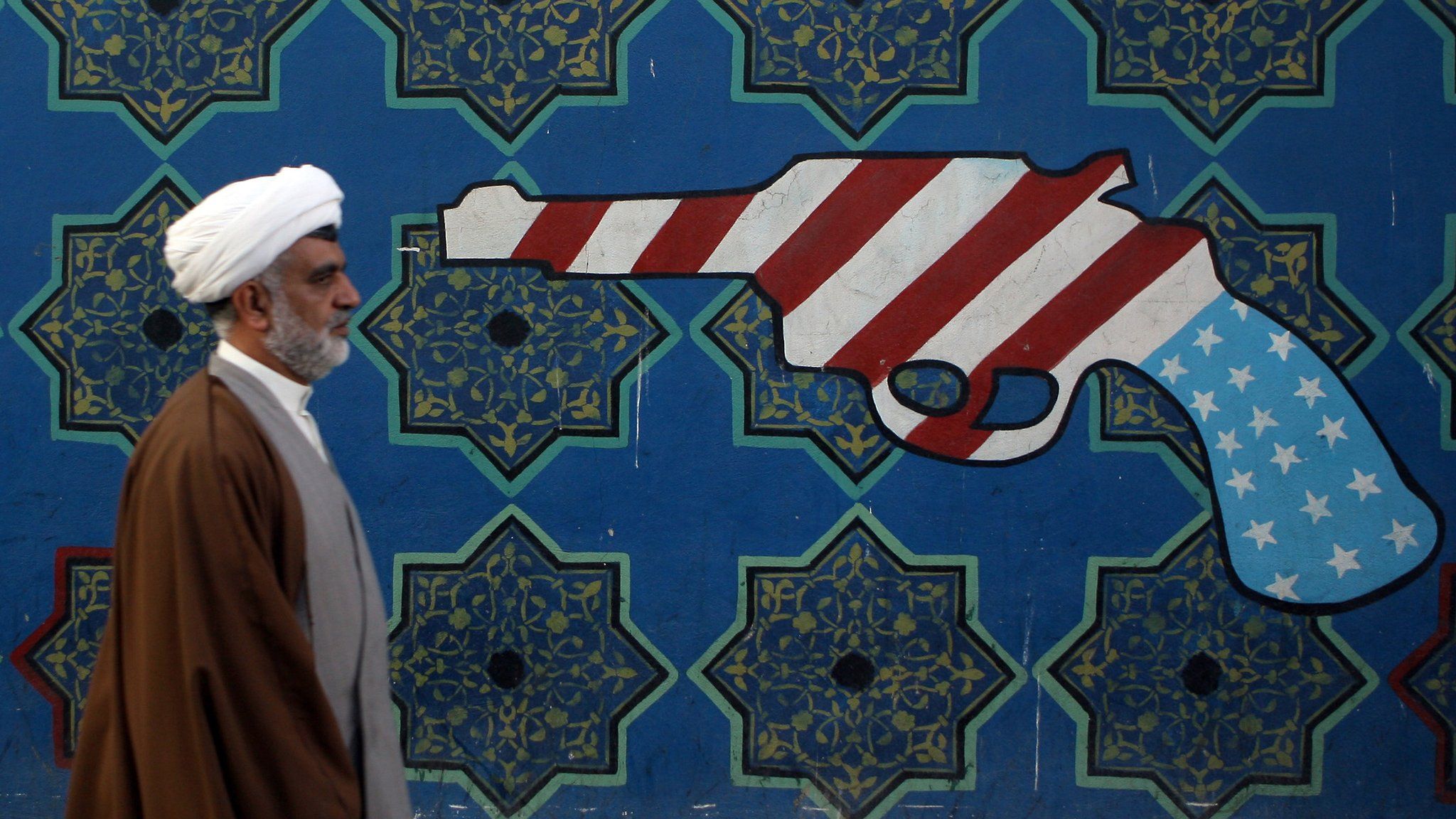 An Iranian clerric walks past a mural on the wall of the former US embassy