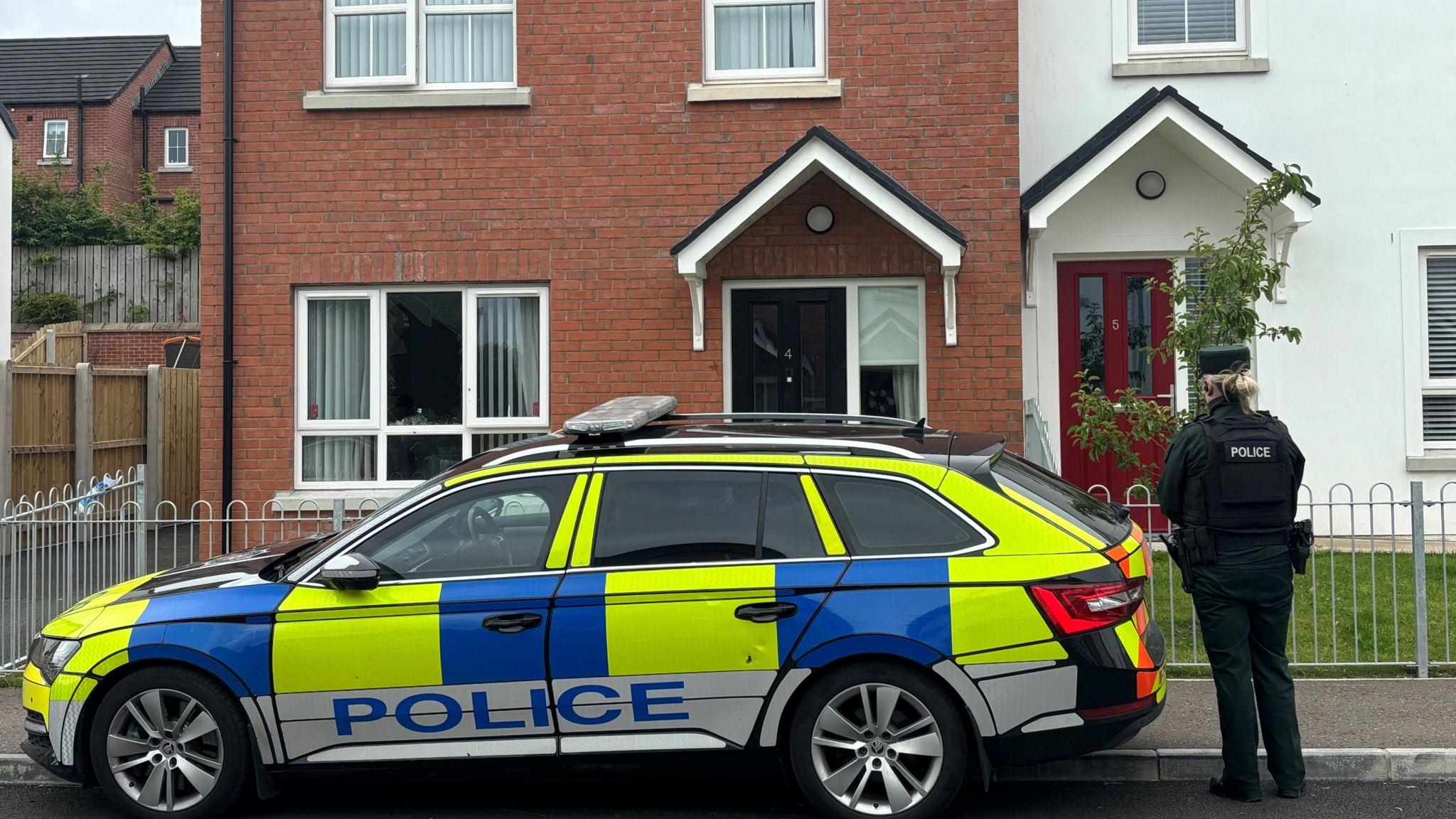 A marked police car is parked outside a red brick house. A uniformed police woman stands with her back to the camera. 