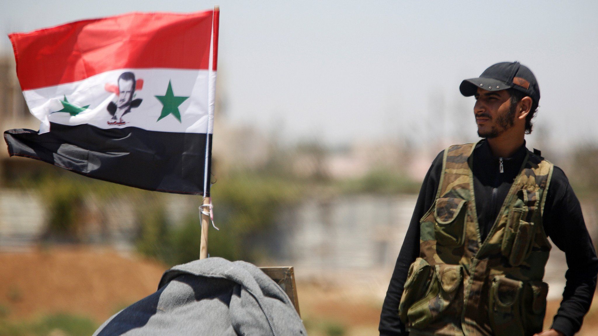A Syrian army soldier stands next to a Syrian flag in Umm al-Mayazen, in the Deraa countryside, Syria (10 July 2018)