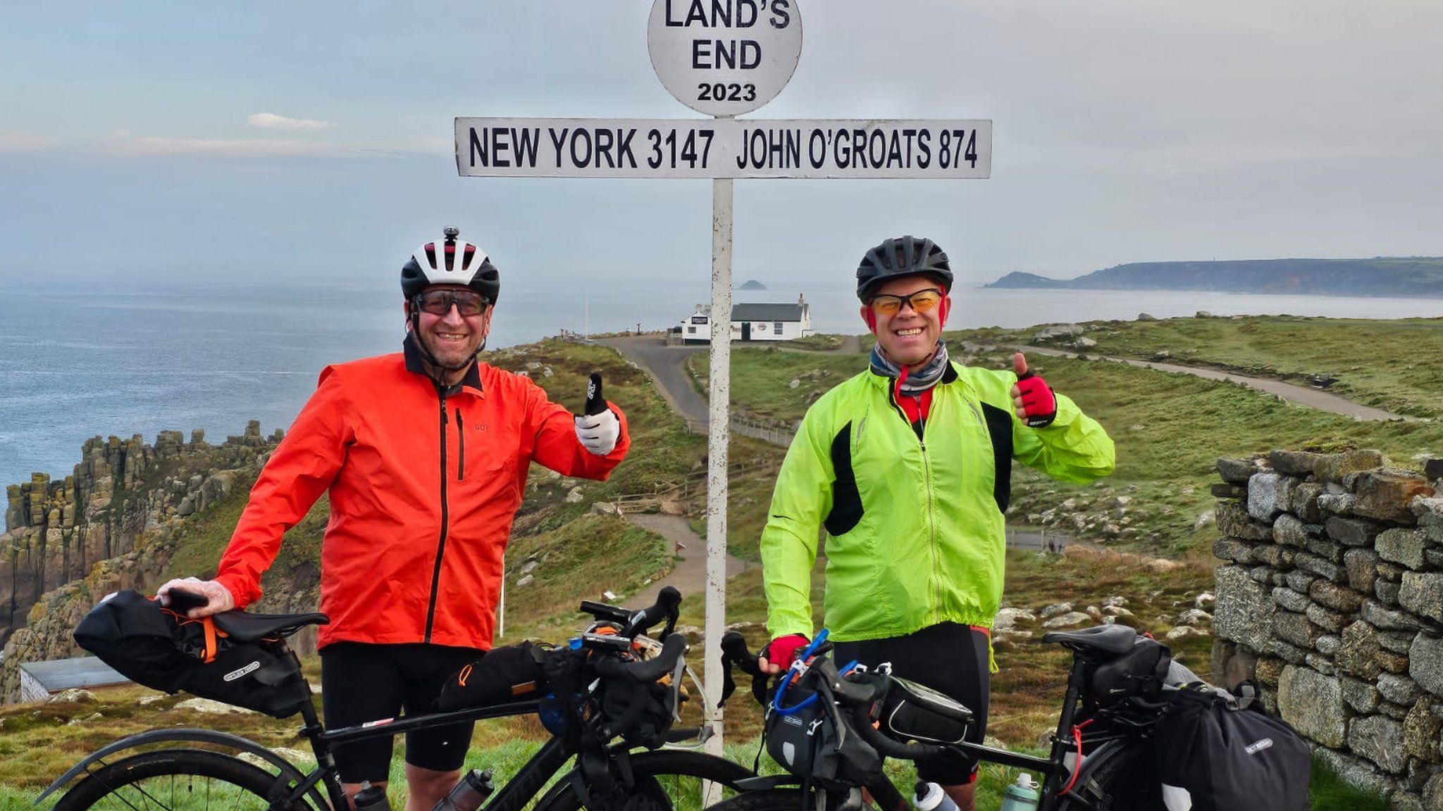 Mark and Ian standing with their bikes in front of the landmark sign at Land's End, Cornwall.