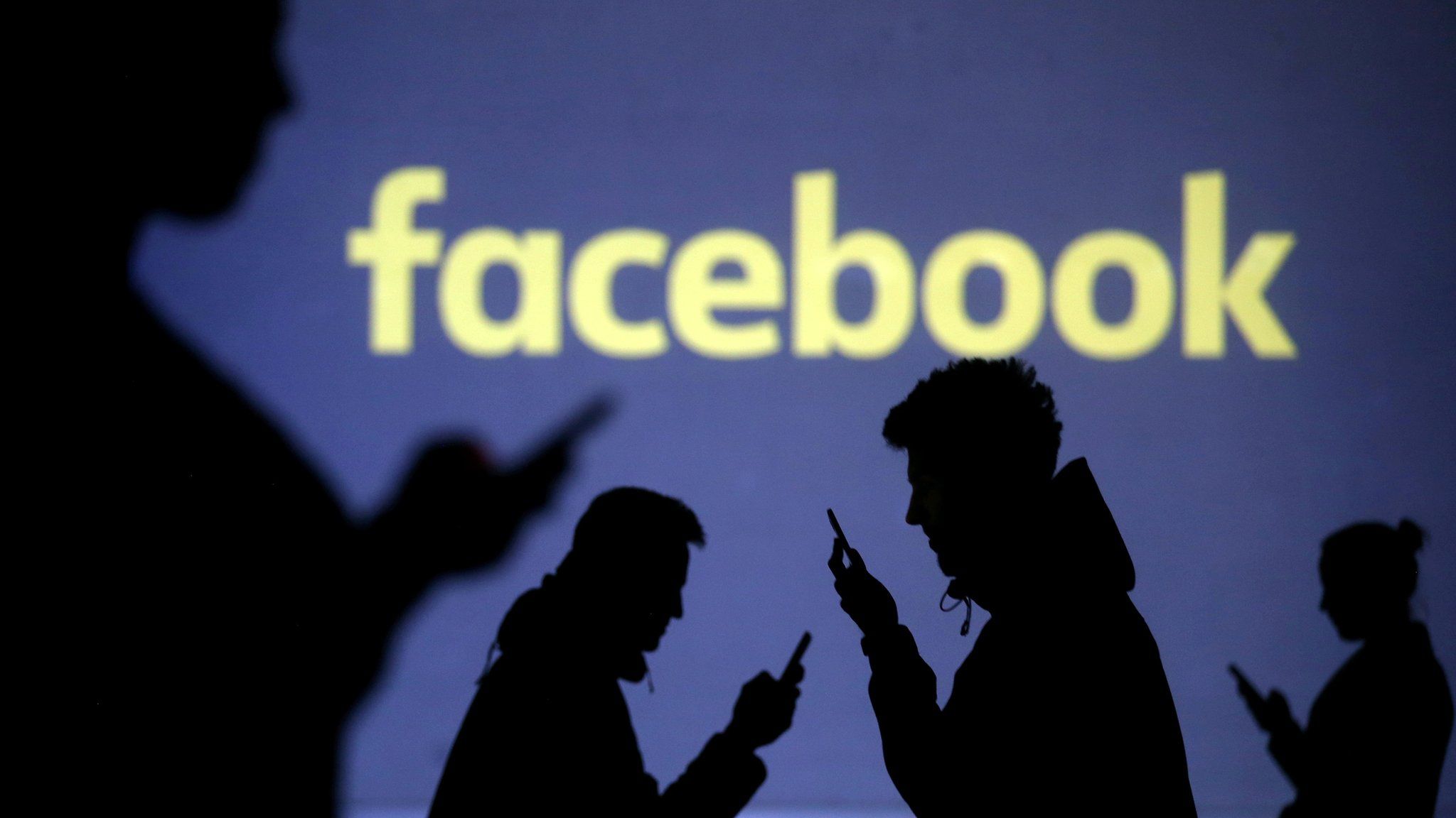 Silhouettes of mobile users are seen next to a screen projection of Facebook logo in this picture illustration taken March 28, 2018.