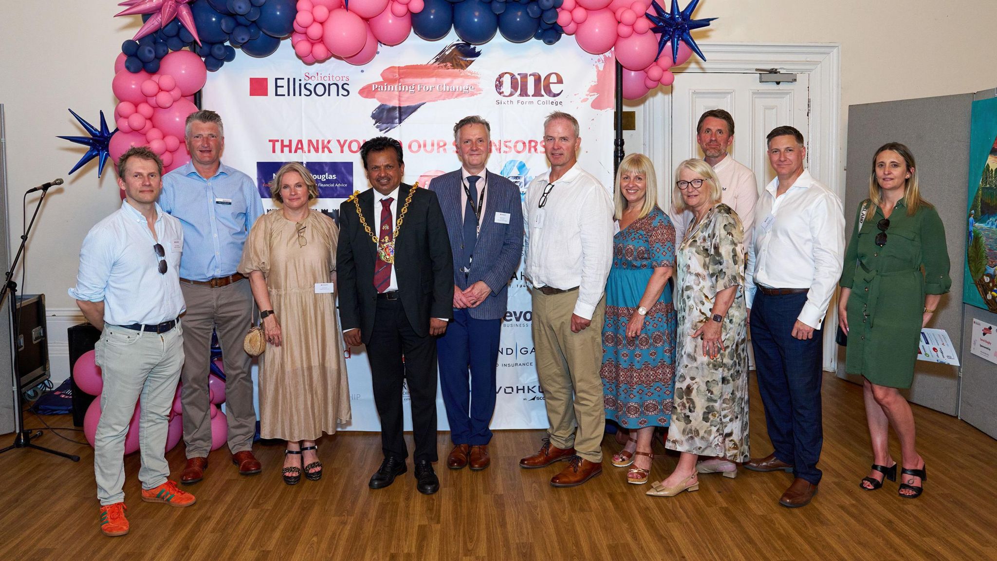Ipswich mayor Elango Elavalakan wearing a suit and mayoral chain standing in a line with workers from Ellisons Solicitors at Ipswich Town Hall. Behind them are pink balloons and a screen that reads "Thank you to our sponsors".