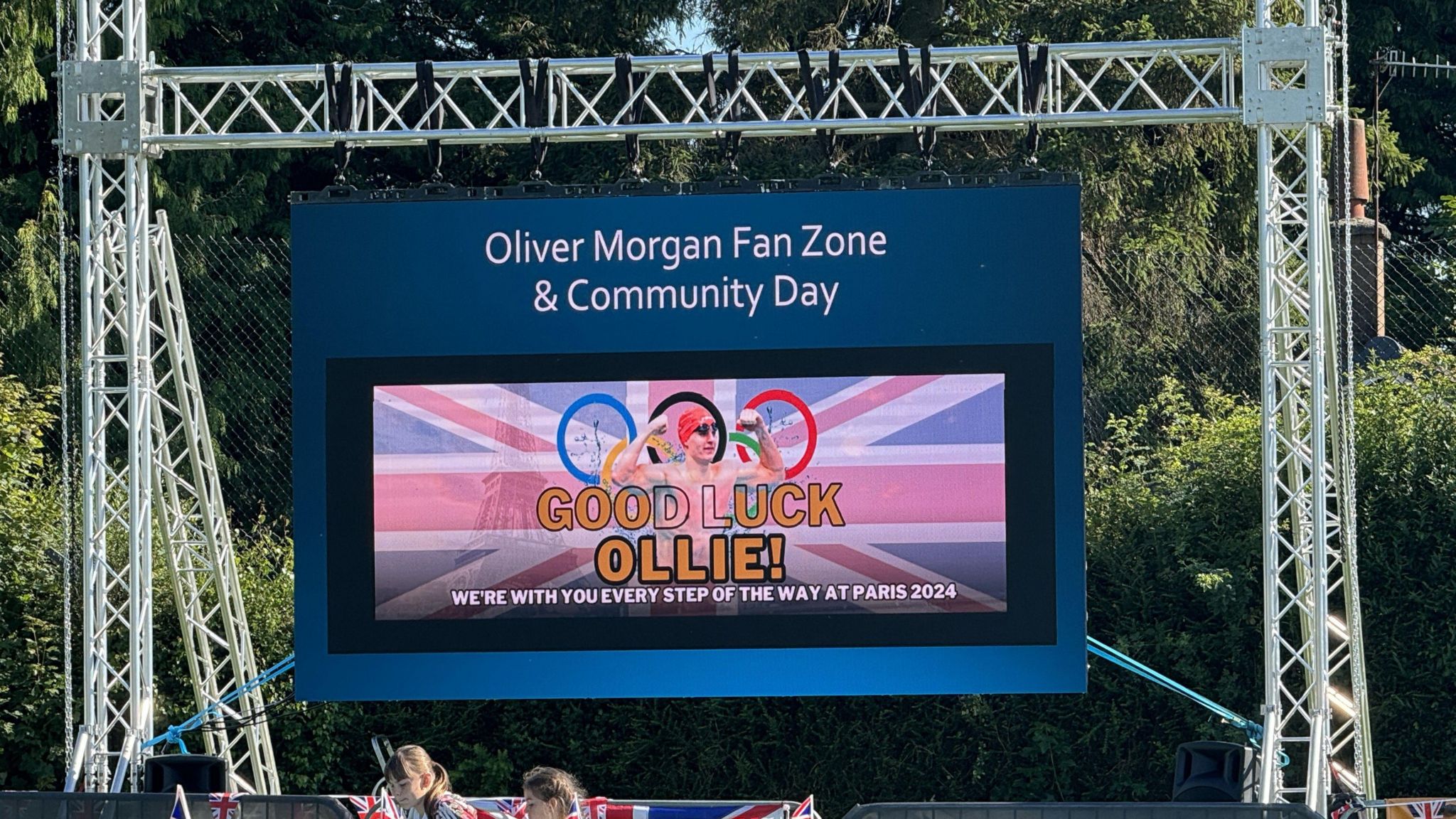 A large screen has been put up on grey meal scaffolding. The screen is blue and reads "Oliver Morgan Fan Zone and Community Day." Below that writing is a photograph of Ollie Morgan within the Olympic rings, and it reads "Good Luck Ollie, we are with you every step of the way at Paris 2024"