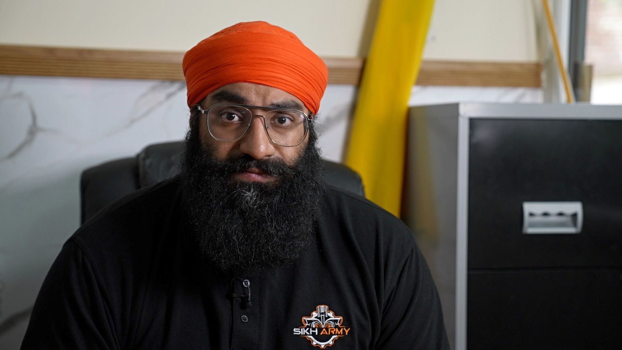 A man with a very full black beard, moustache, thin framed silver glasses and an orange turban. He is wearing a black jacket with an emblem that says Sikh Army.