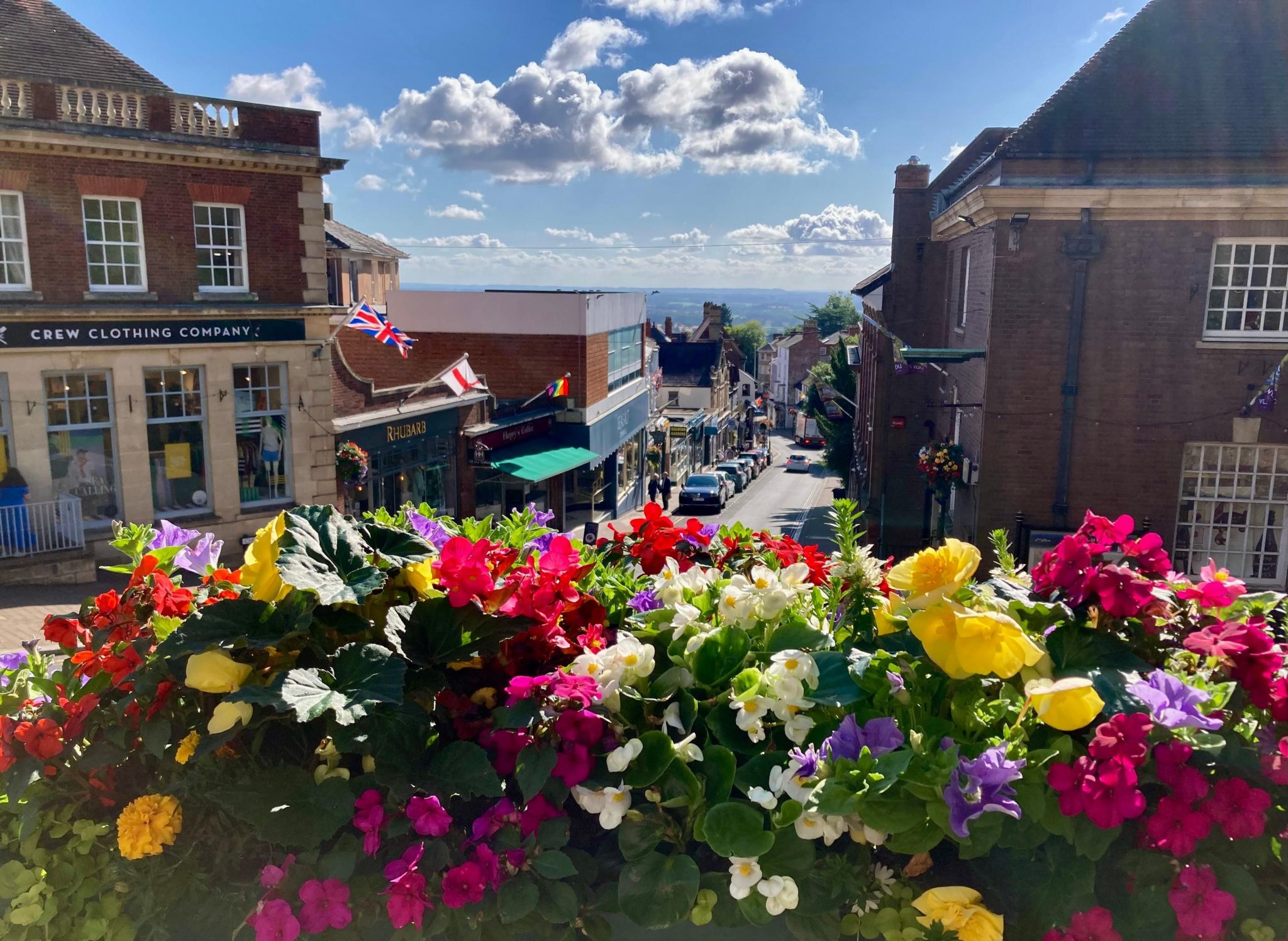 A mix of flowers in red, pink, yellow, white and purple in a planter in the foreground, with a view of Great Malvern town centre behind 