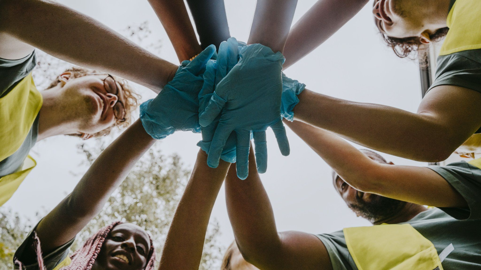 A group of people in high vis jackets with blue plastic gloved hands stand in a circle with their hands together in a show of teamwork