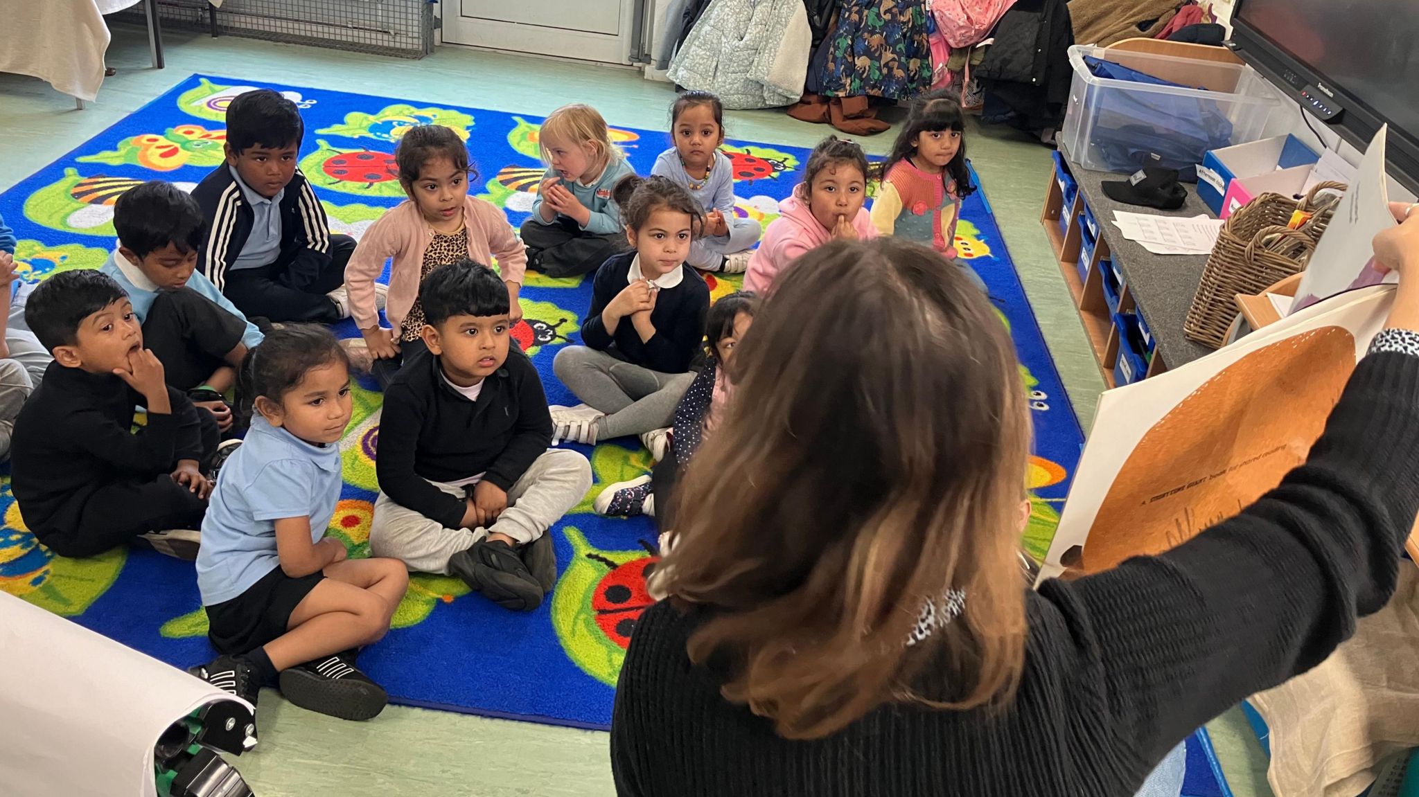 A teacher with brown hair holds up a large book to a class of pre-schoolers sitting on a colourful mat with a bug pattern, listening to the story. Many are sitting with their legs crossed with their hands together. 