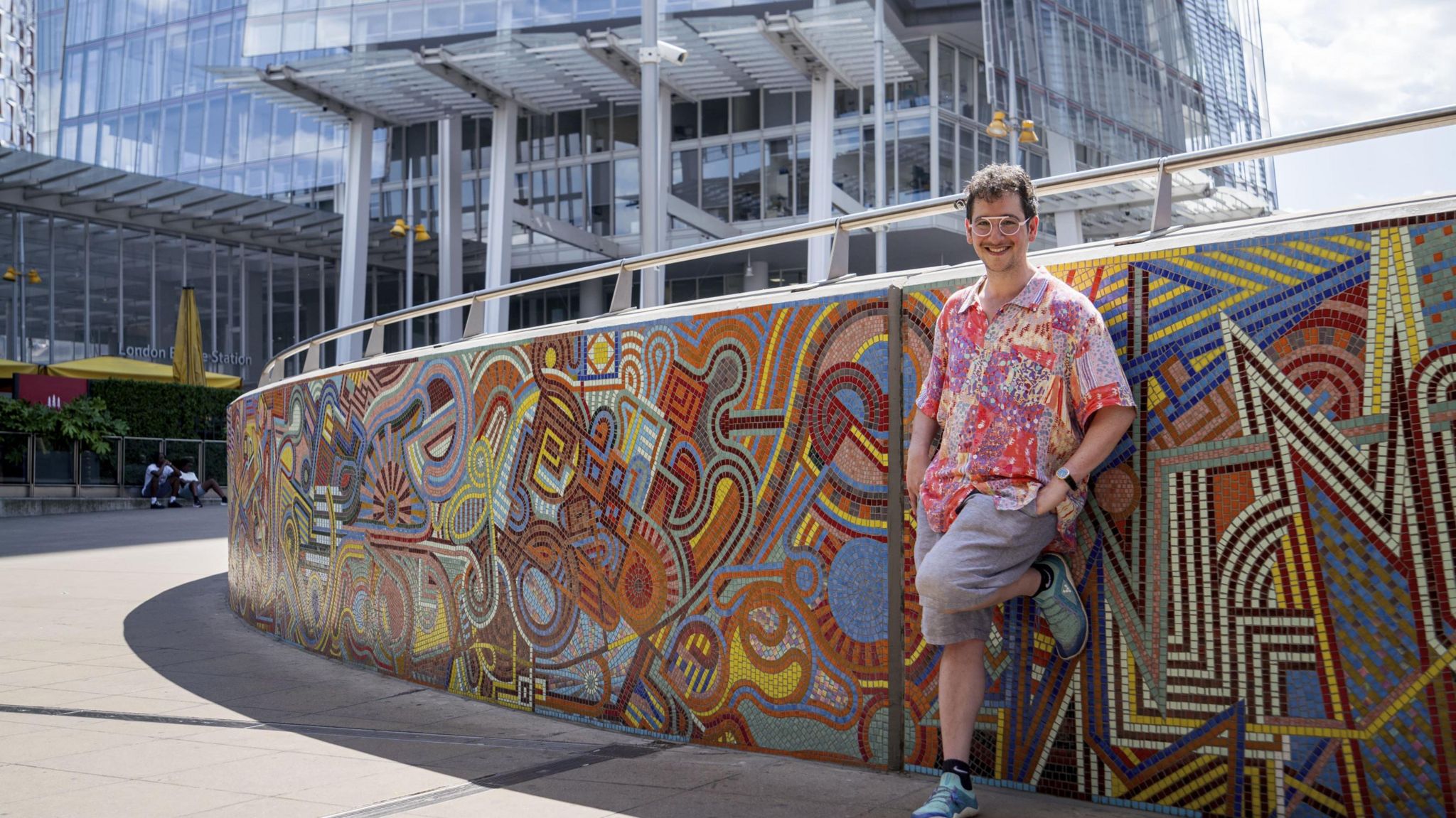 Adam Nathaniel Furman in front of the mural, standing with one leg up against the mural and wearing a colourful shirt, smiling at the camera