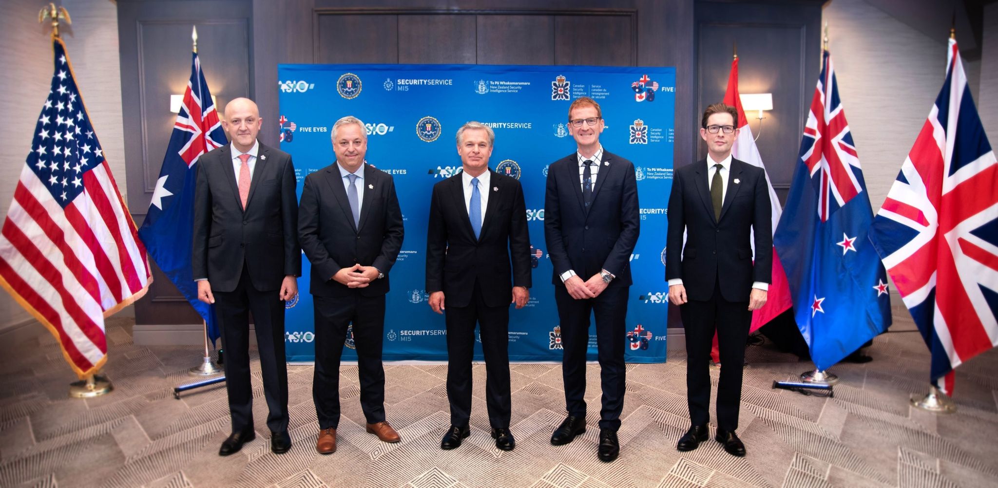 The heads of US, UK, Australian, Canadian and New Zealand security agencies stand in a row flanked by flags.