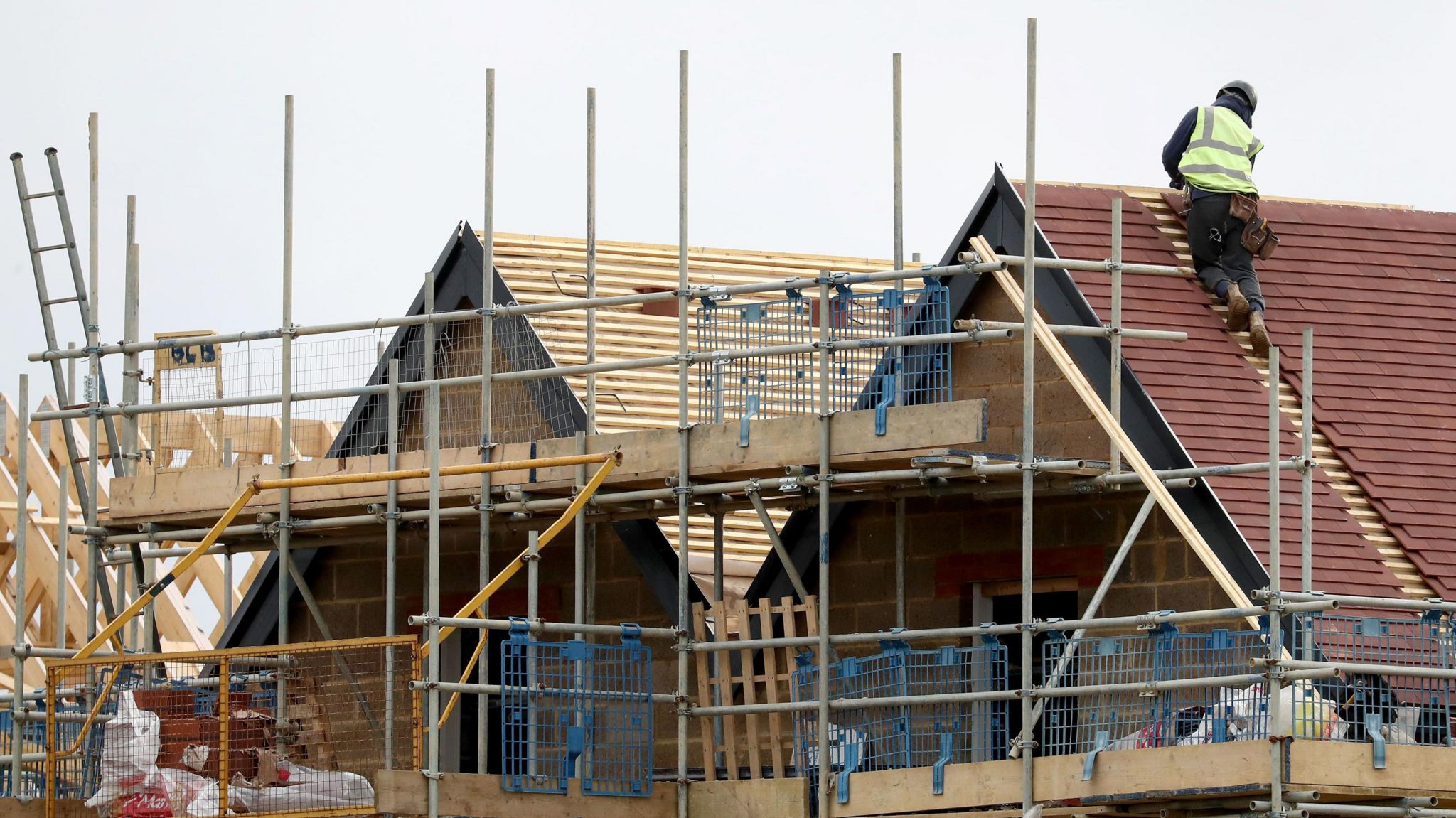 A builder wearing a yellow high-visibility jacket on the red-tiled roof of a house under construction. In the foreground is scaffolding and a tall ladder.