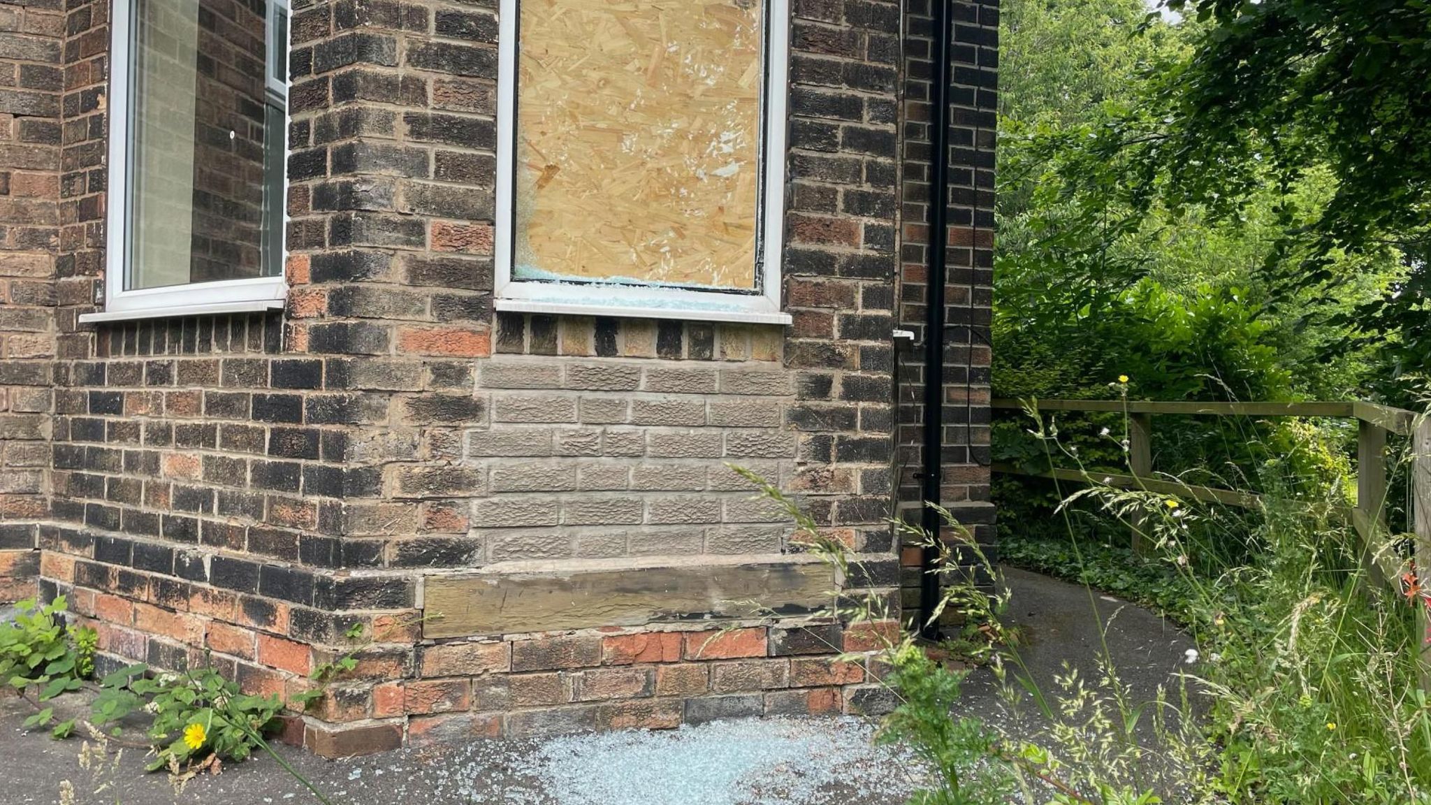 A window is boarded up with broken glass scatted on the ground below