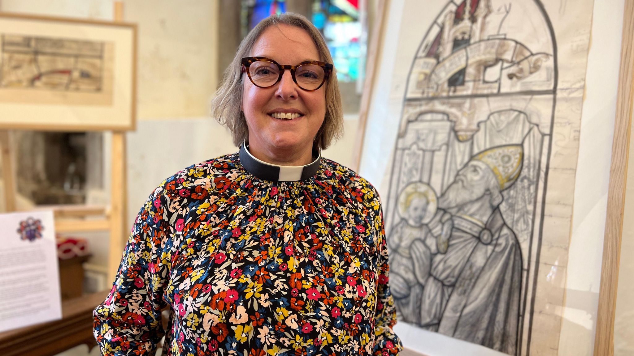 Reverend Allan smiles at the camera with a framed cartoon drawing behind her