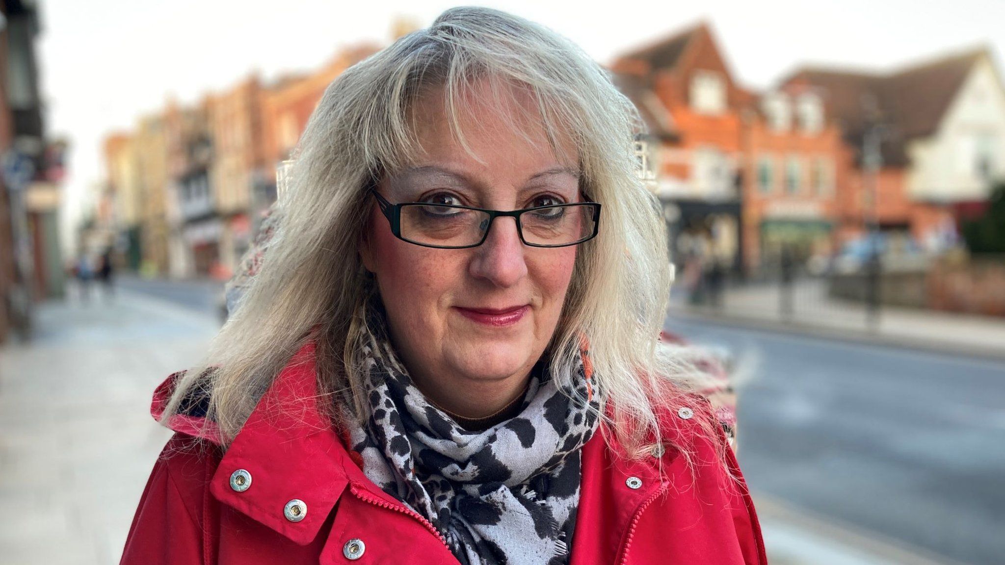 A blonde woman smiling. She is wearing glasses and a red coat.