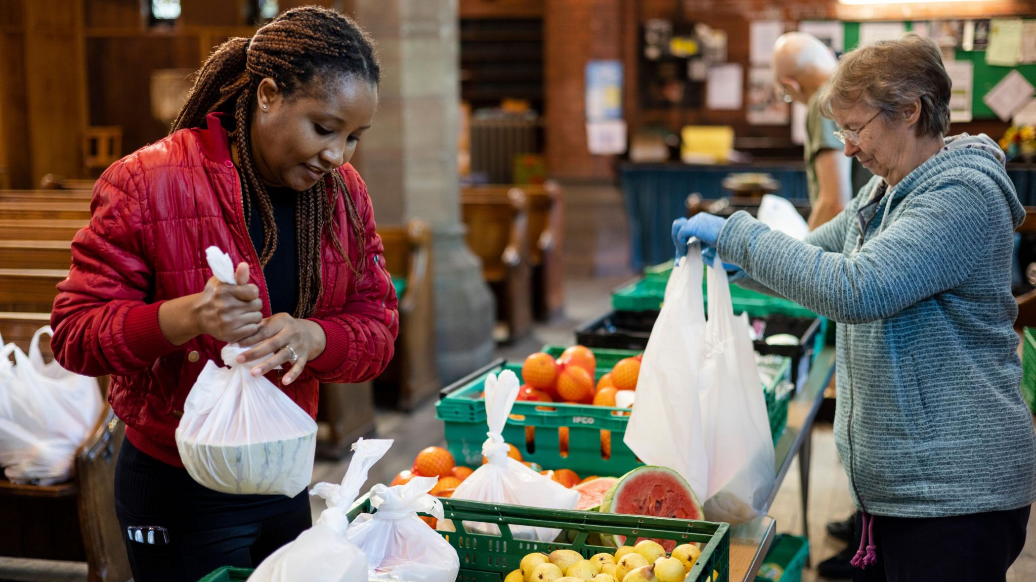 A woman with dreadlocks, red jacket and black trousers and holding a white carrier bag with a water melon inside looks at a selection of fruit on tables at a food hub. A woman wearing a pale blue zip-up hubby and blue gloves is serving. The food on show includes melons, lemons and oranges and the setting is inside a church